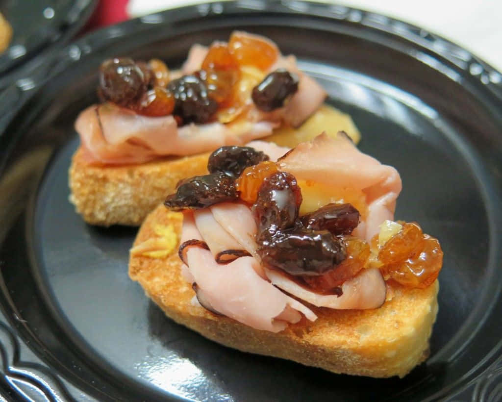 Black Forest Ham 桌布 壁紙 — 兩塊多士麵包，上面放有火腿、提子乾和糖漬橙皮，放在深色碟上。