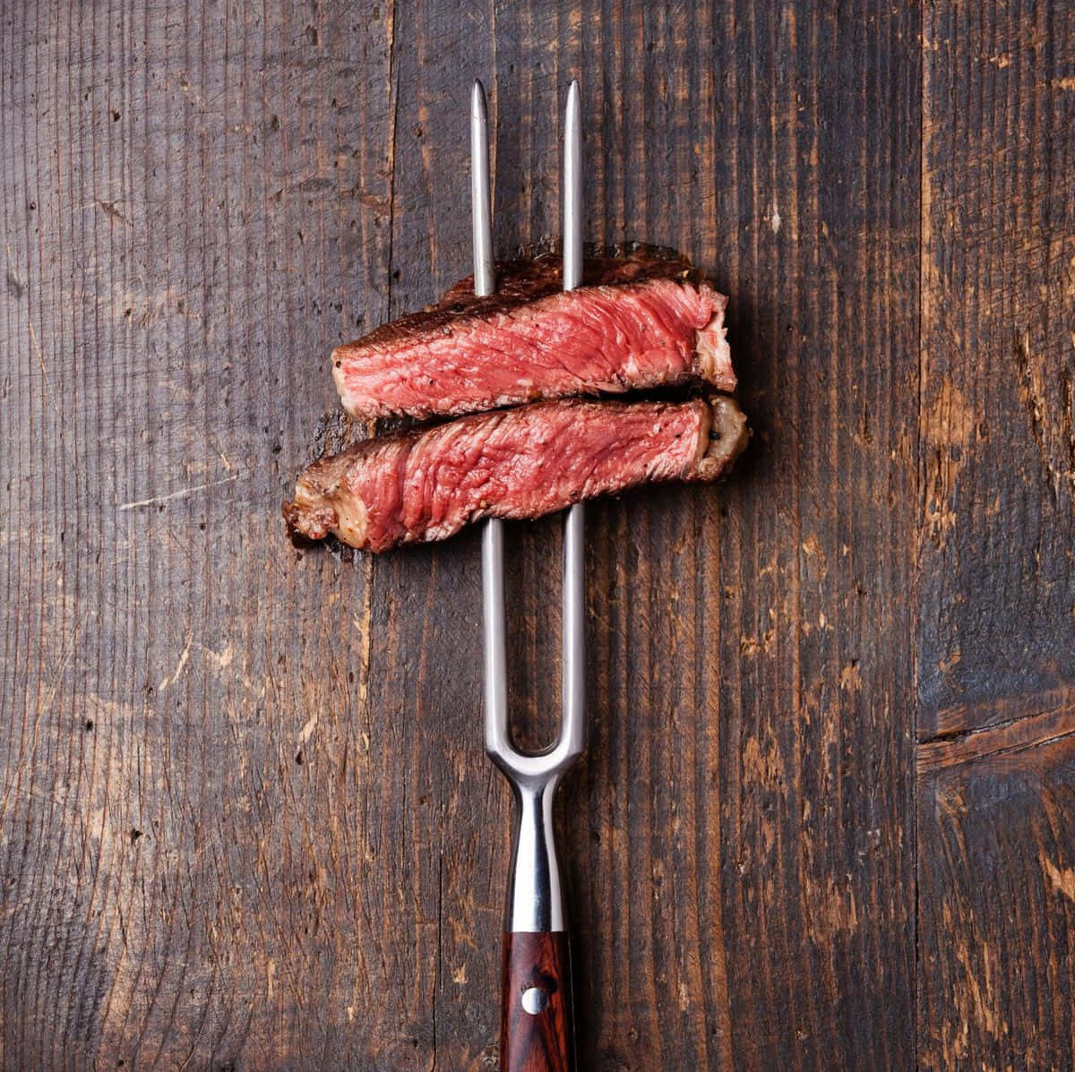 Red Meat 桌布 壁紙 — 兩塊多汁嘅 medium rare 牛扒片用金屬叉叉住，放喺深色鄉村風木質表面上。