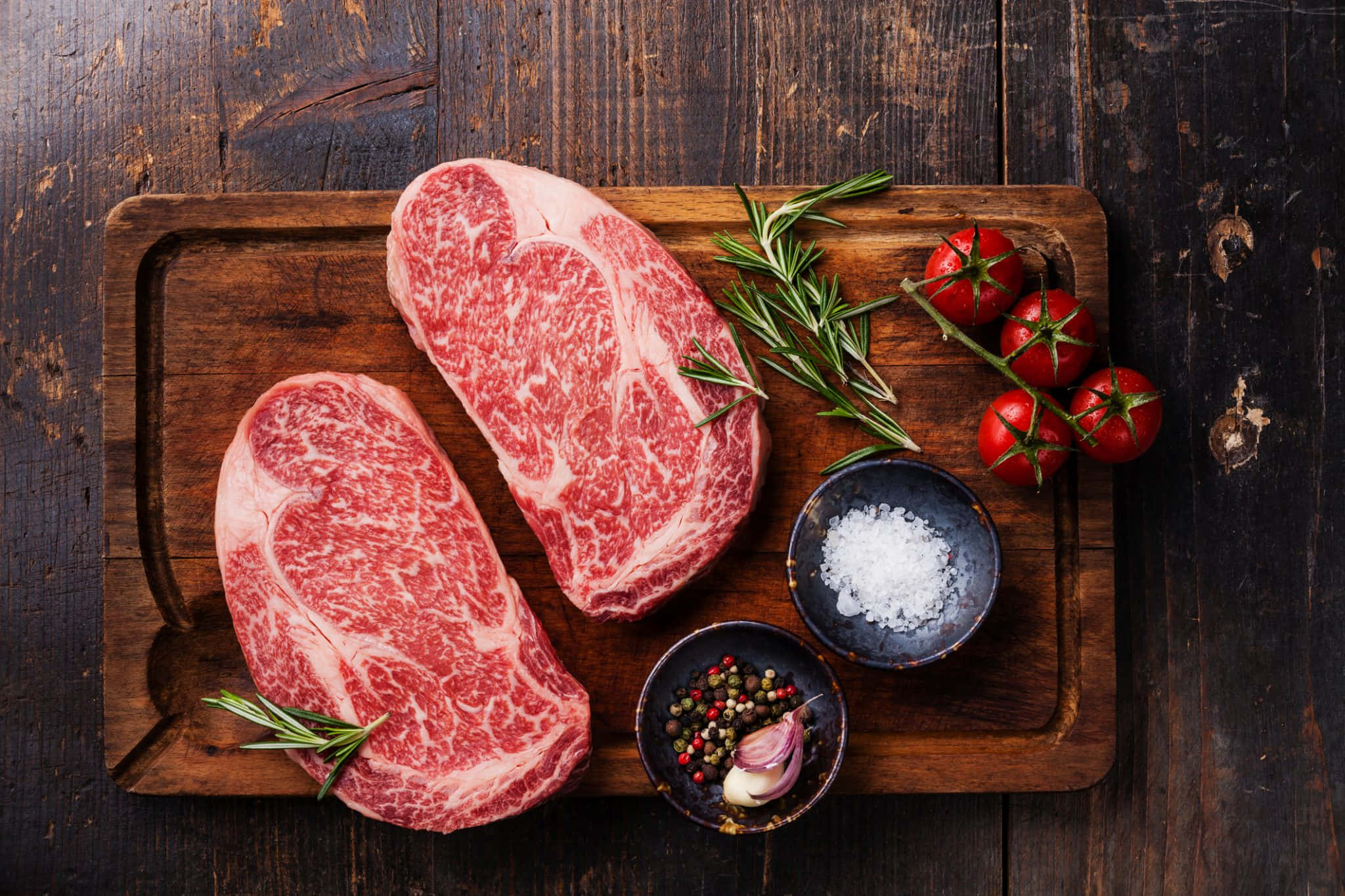 meat 桌布 壁紙 — 兩塊大理石紋牛扒放在質樸的木板上，配以迷迭香、車厘茄、鹽和胡椒粒。