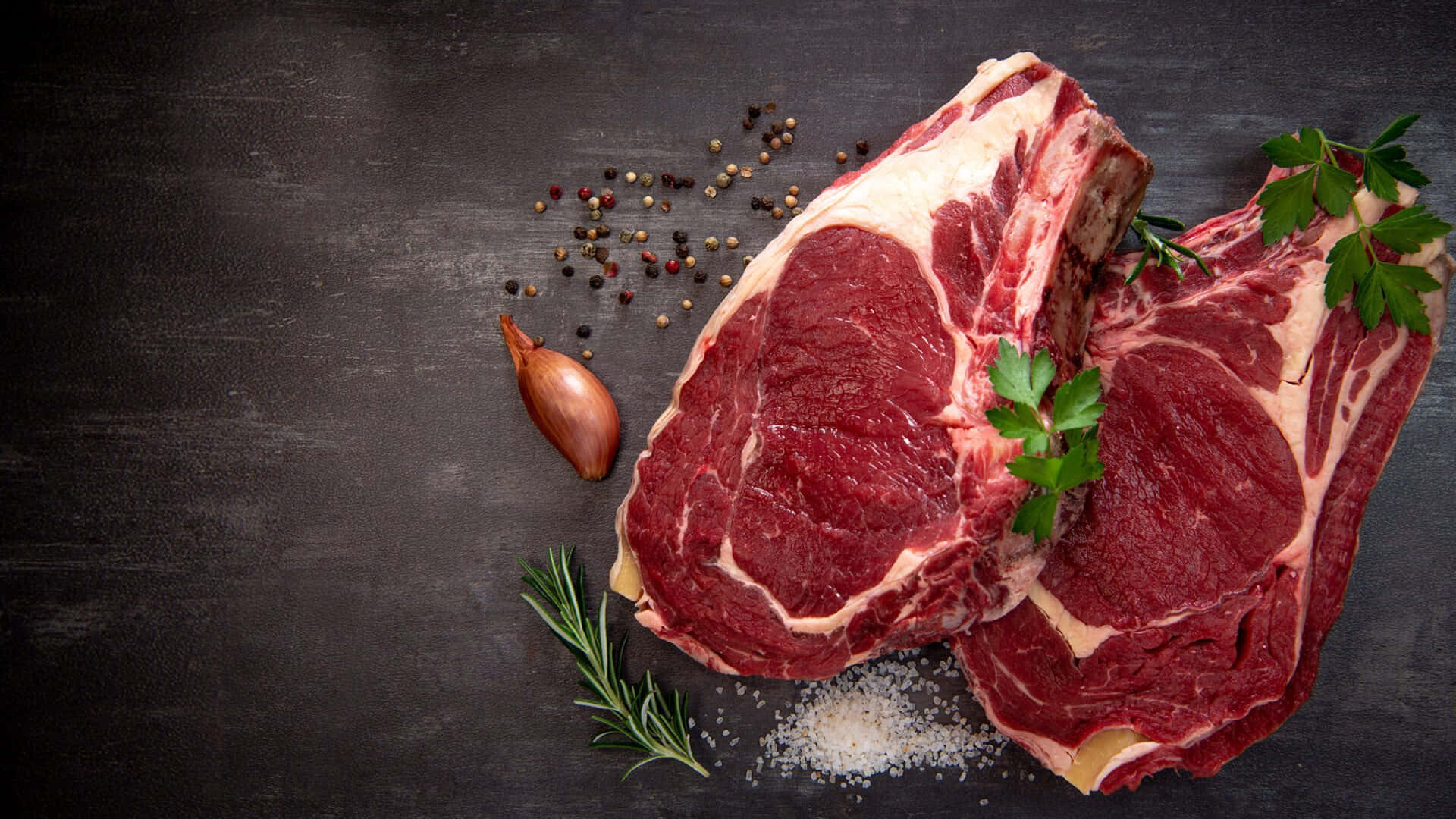 Red Meat 桌布 壁紙 — 兩塊帶骨生肉眼牛排，以迷迭香、歐芹、鹽和胡椒粒裝飾，放在深色質樸的表面上。