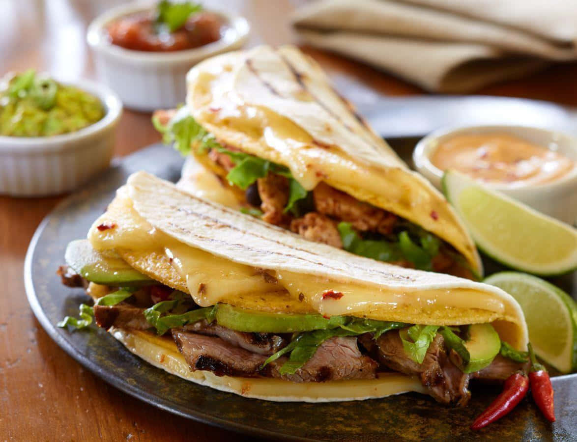 Taco 桌布 壁紙 — 兩塊烤雞肉玉米餅，配上融化芝士、牛油果和生菜，放在深色碟上，伴以青檸角和蘸醬。