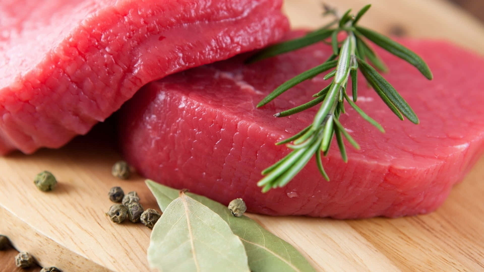 Red Meat 桌布 壁紙 — 兩塊生牛肉扒放在木板上，以迷迭香、月桂葉和胡椒粒裝飾。