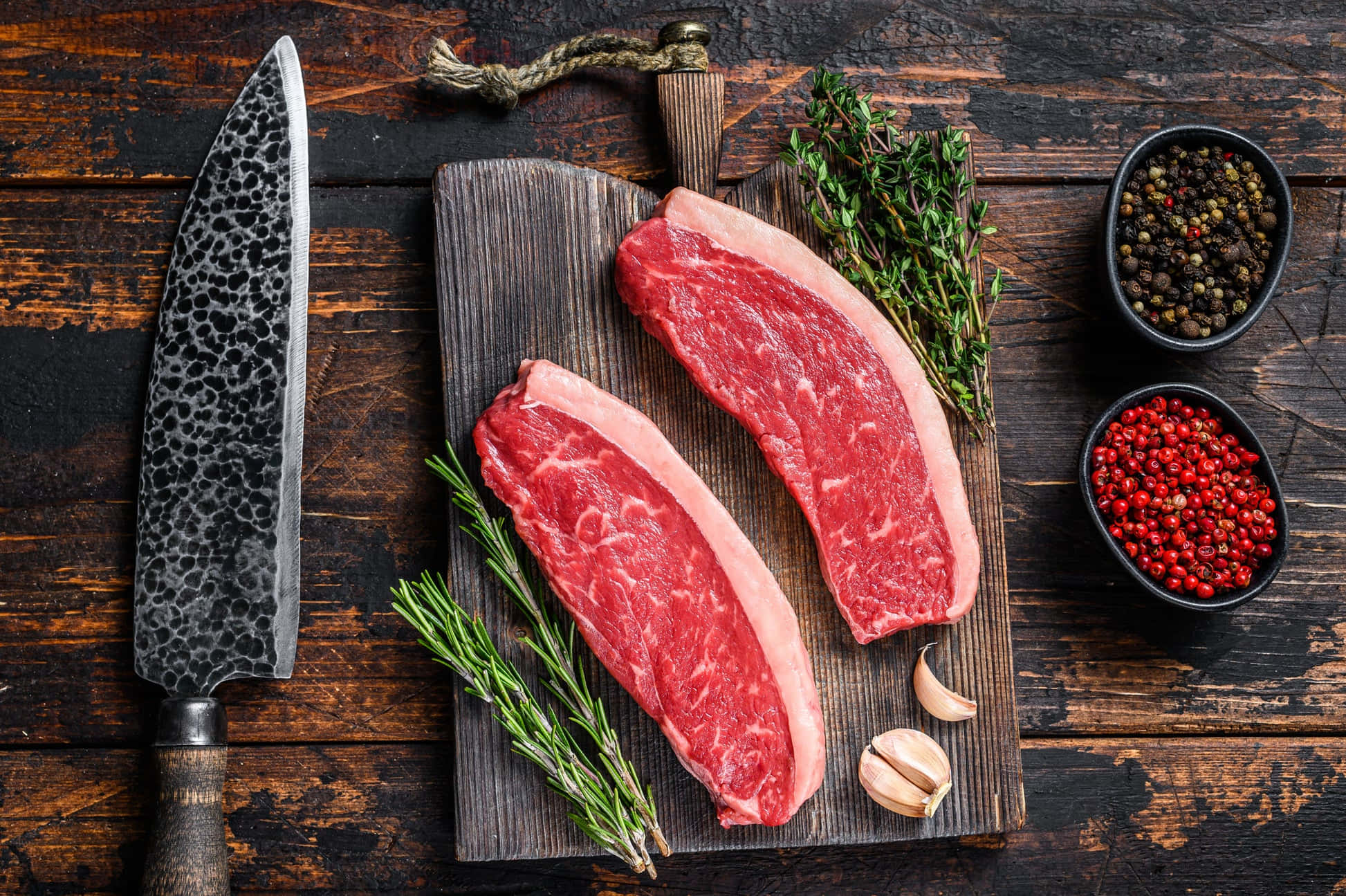 Red Meat 桌布 壁紙 — 兩塊生牛肉扒放在木砧板上，旁邊是新鮮香草、大蒜、胡椒粒和一把錘紋刀，置於質樸的深色木質表面上。