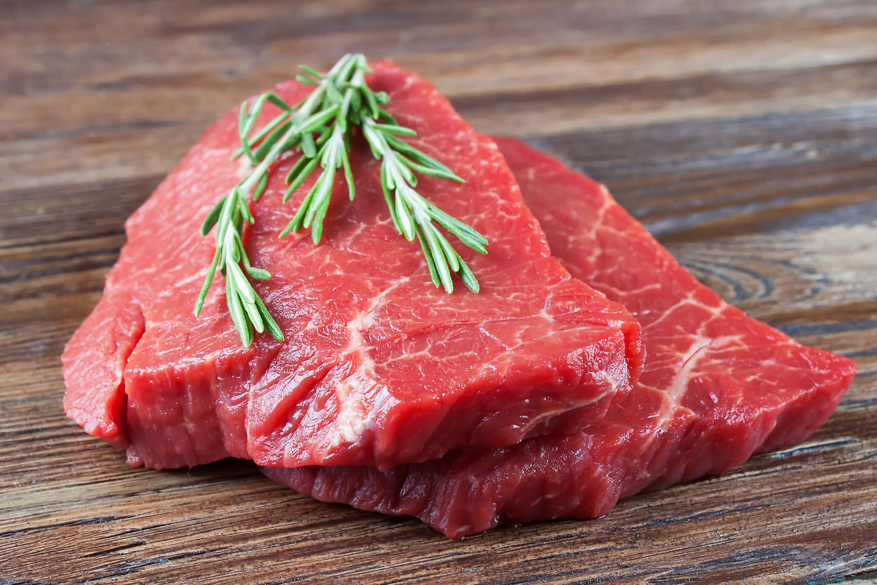 Red Meat 桌布 壁紙 — 兩塊生牛肉扒配迷迭香枝條，放在質樸的木質表面上。