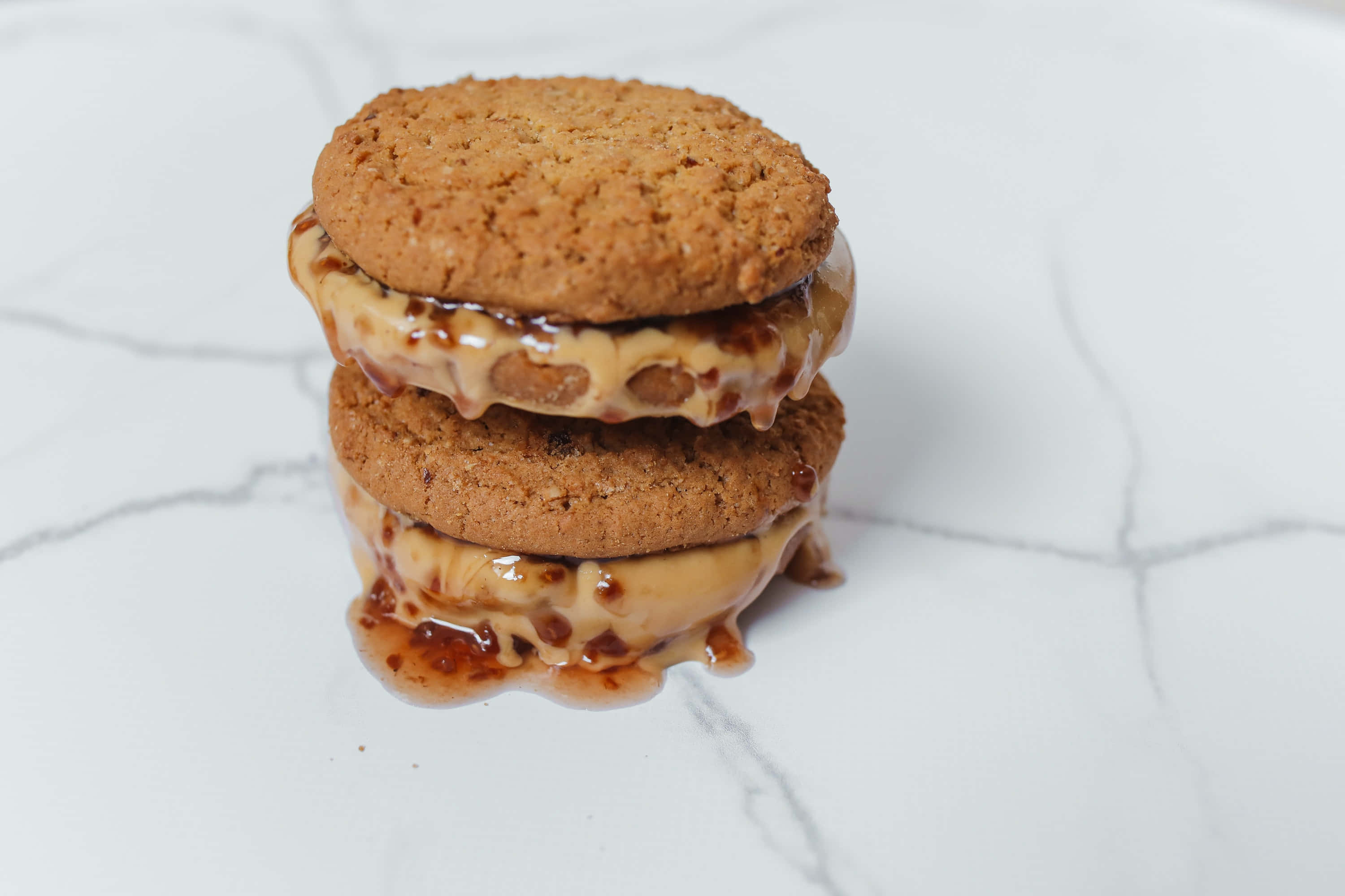 Caramel 桌布 壁紙 — 兩塊疊起的燕麥曲奇，焦糖餡料滲落到白色大理石表面上。