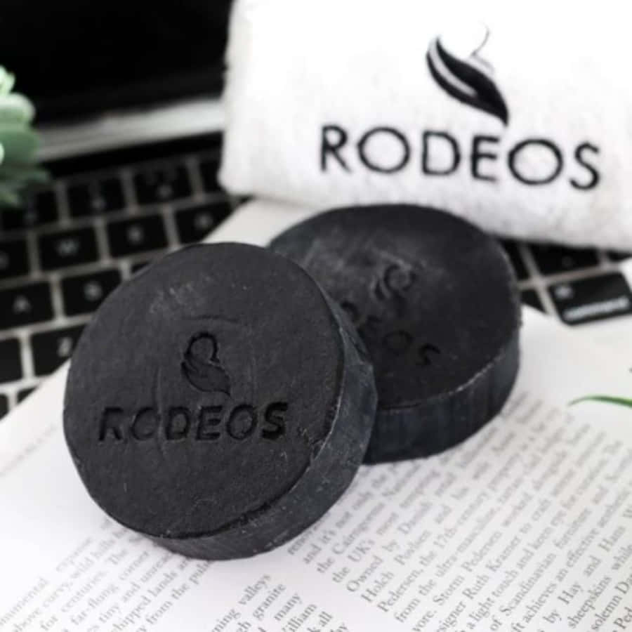 Black Soap 桌布 壁紙 — 兩塊磨砂黑色圓形RODEOS肥皂放在一本打開的書上，旁邊有品牌毛巾和手提電腦鍵盤。