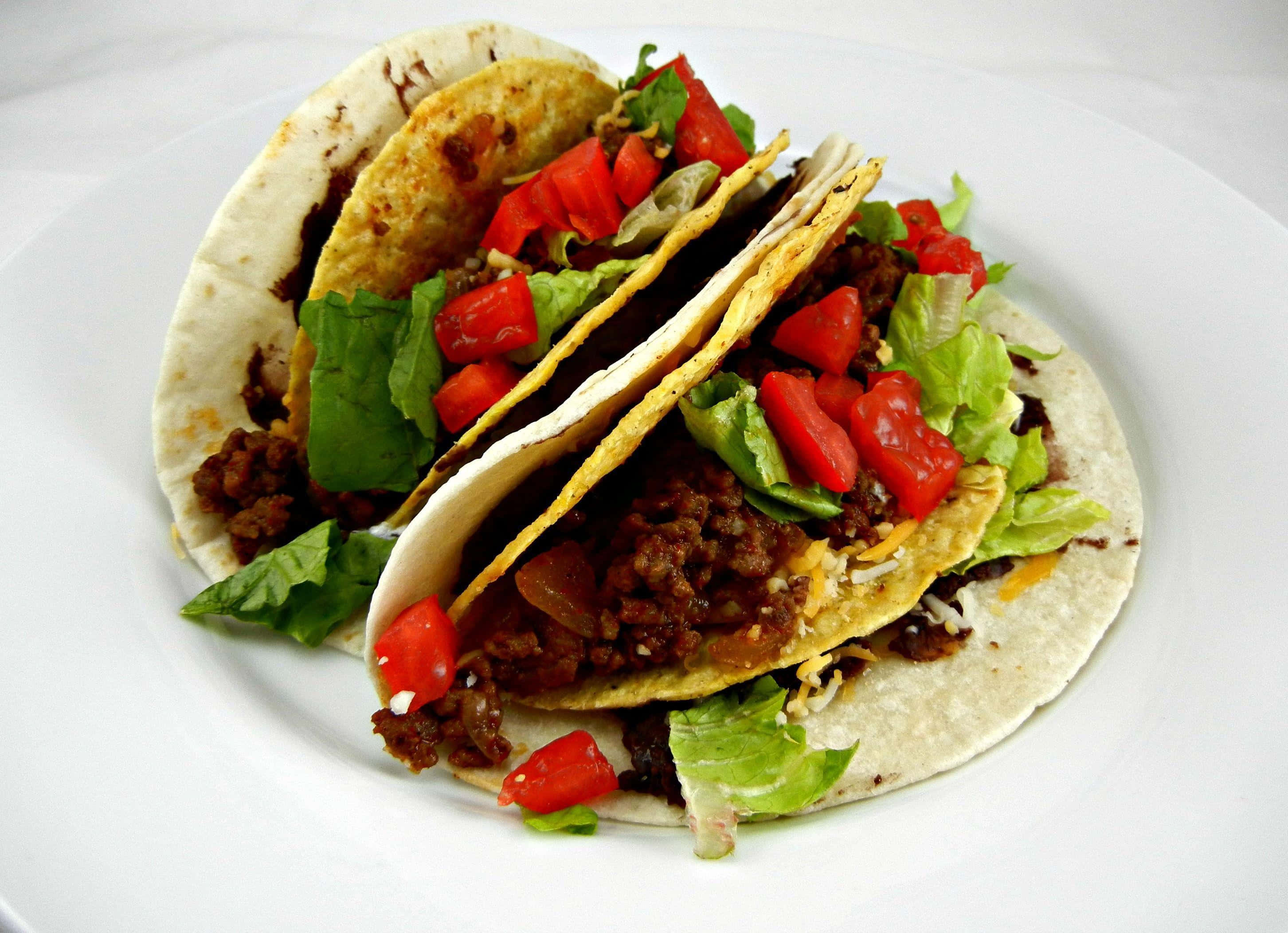 Taco 桌布 壁紙 — 兩塊脆皮墨西哥卷餅，內餡有調味肉碎、生菜和番茄粒，誘人地放在白色碟上。