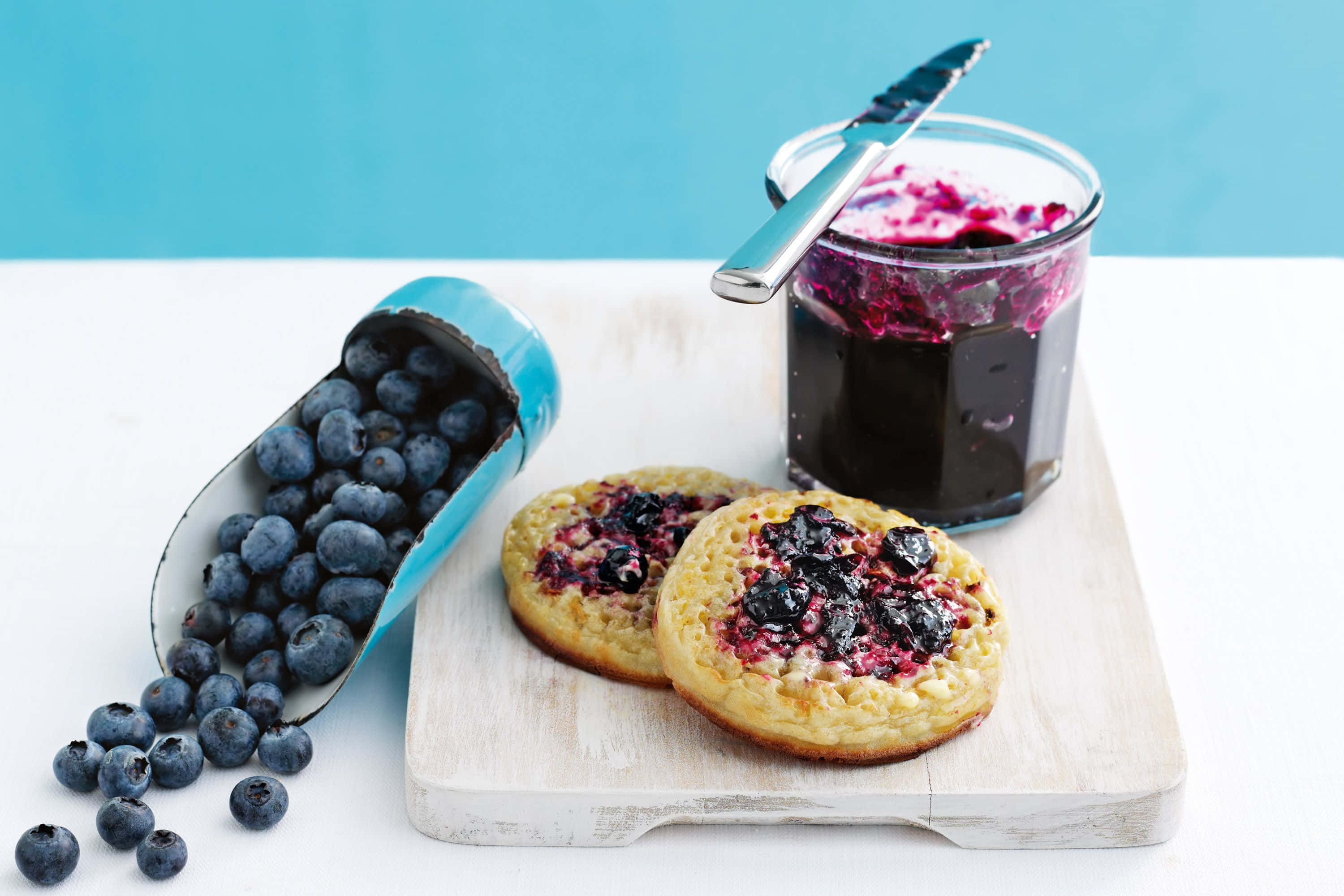 Blueberry Jam 桌布 壁紙 — 兩塊金黃色的藍莓煎餅，上面鋪滿新鮮莓果和果醬，放在木板上，旁邊有一個翻倒的藍莓容器和玻璃罐。