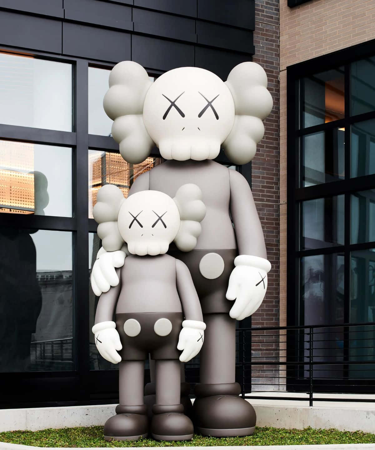 Kaws Figures 桌布 壁紙 — 兩尊巨型KAWS Companion雕像並排站立在一棟現代建築外，建築有玻璃窗和磚牆。