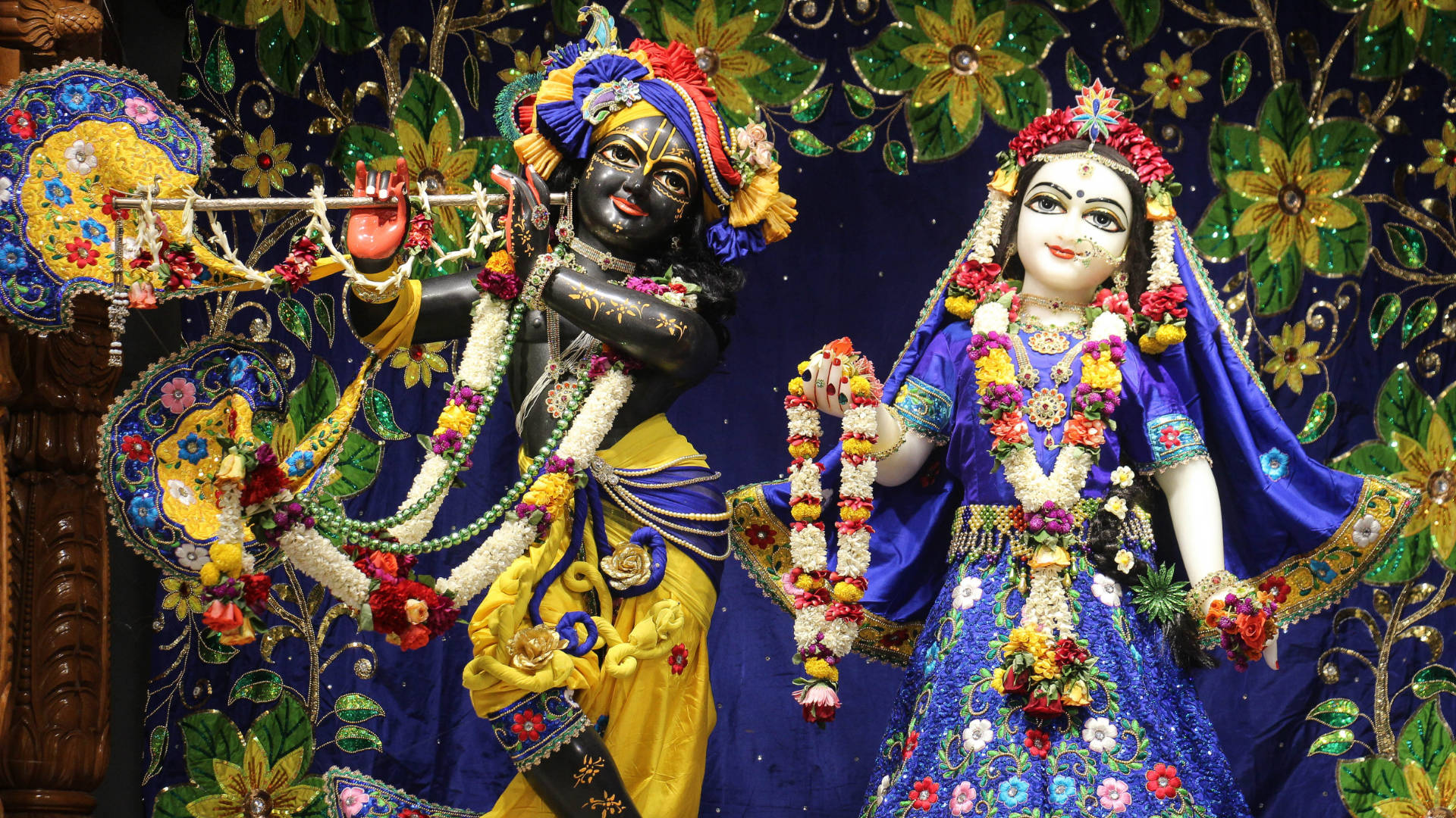 Krishna Desktop 桌布 壁紙 — 兩尊精心裝飾的印度教神祇克里希納和拉妲，身披鮮豔花環和華麗刺繡服飾，立於珠寶花卉背景前。