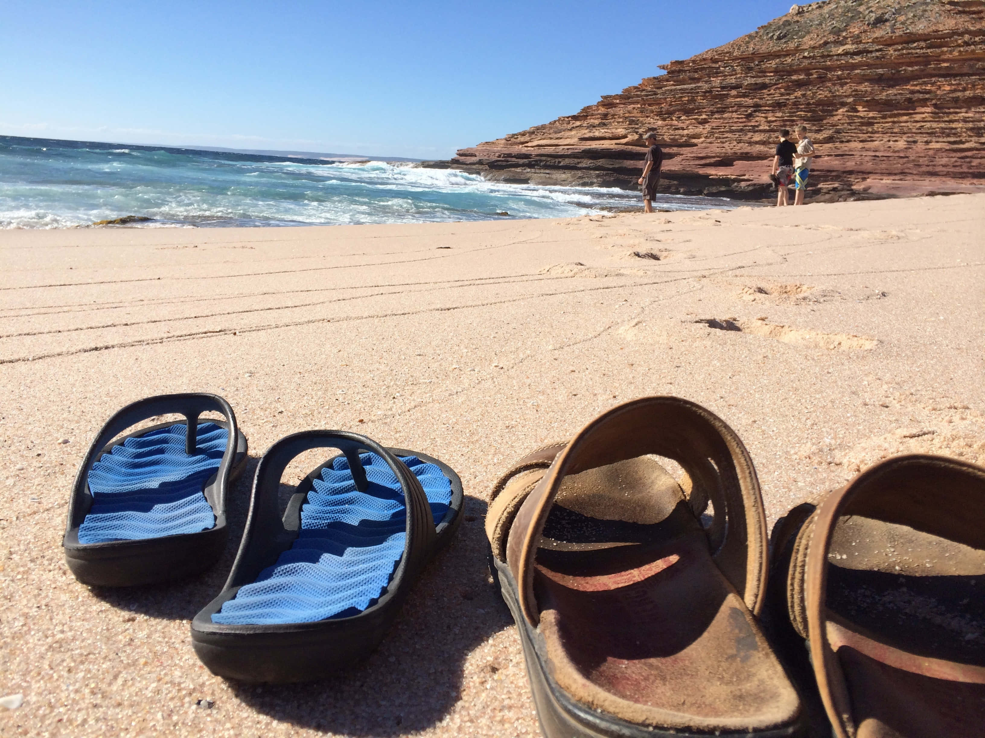 Beach Sandals 桌布 壁紙 — 兩對涼鞋放在陽光普照的沙灘上，海浪拍打著紅色懸崖，遠處有人走動。