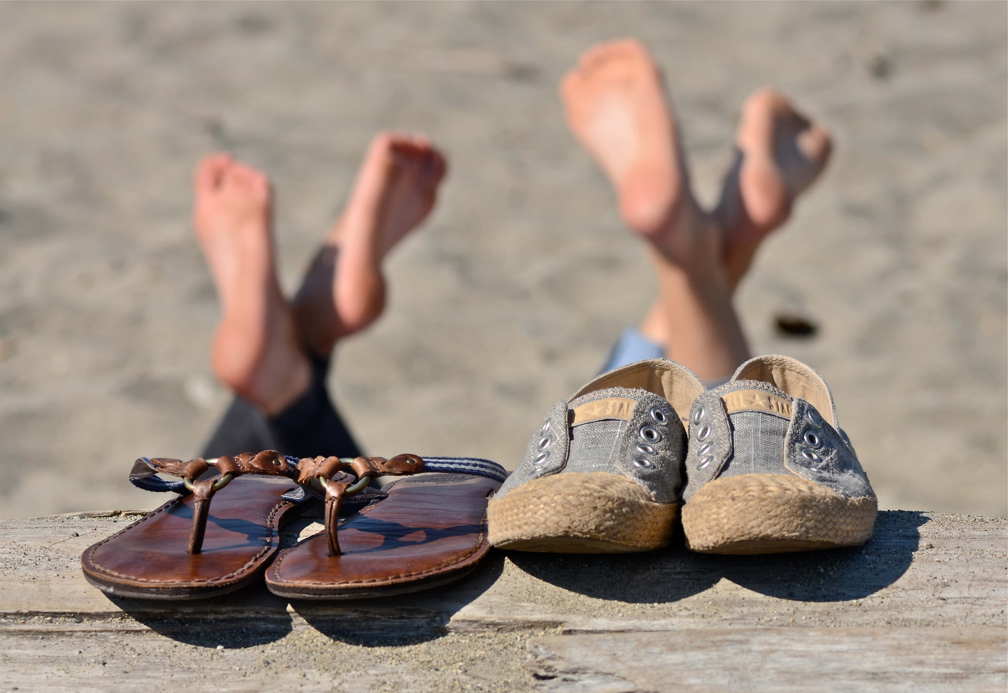 Beach Sandals 桌布 壁紙 — 兩對鞋放在一塊木板上，赤腳在陽光明媚的海灘上踢起沙子。
