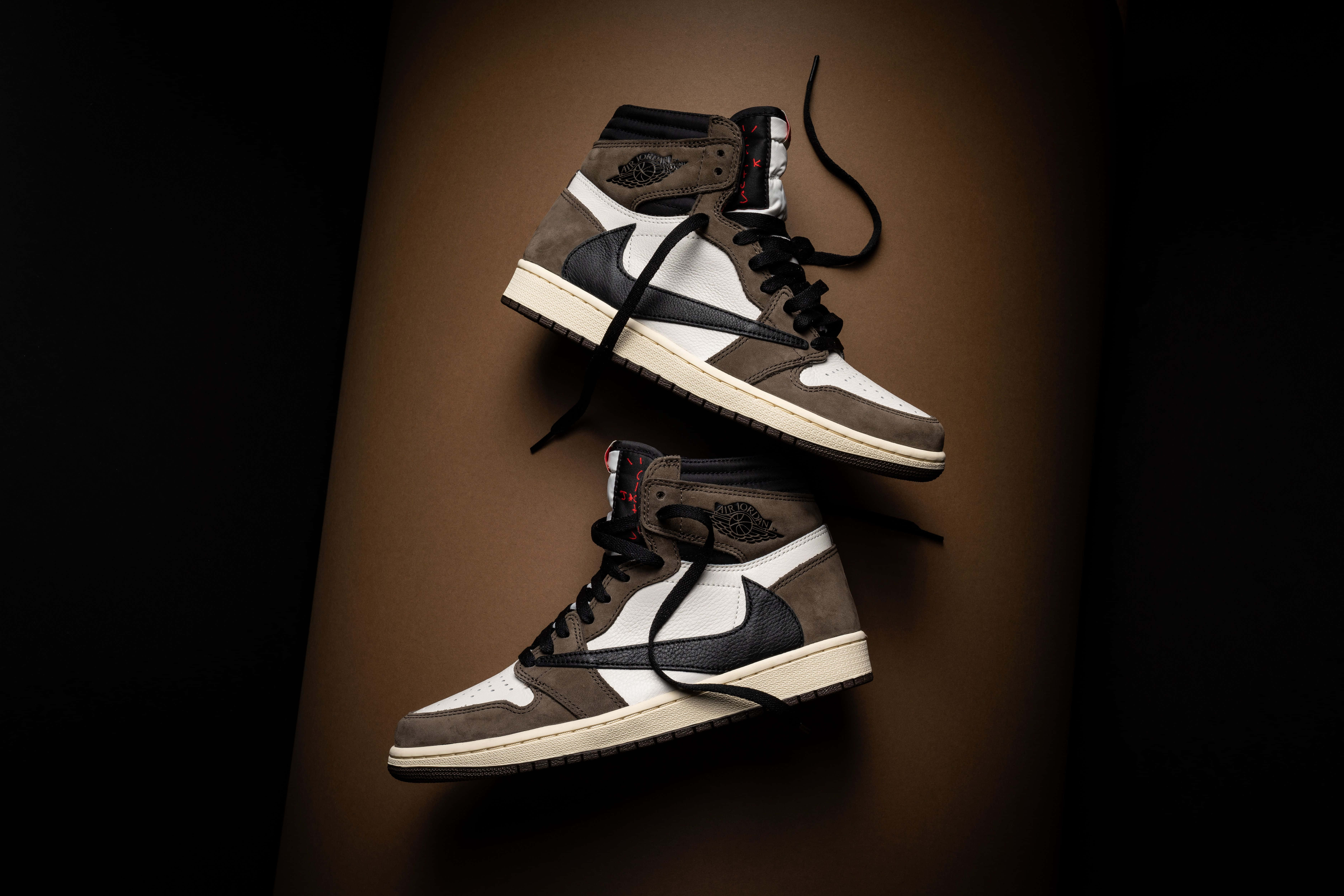 Travis Scott Jordan 1 桌布 壁紙 — 兩對高筒運動鞋，帶有棕色、白色和黑色點綴，在深棕色背景上以戲劇性燈光漂浮。