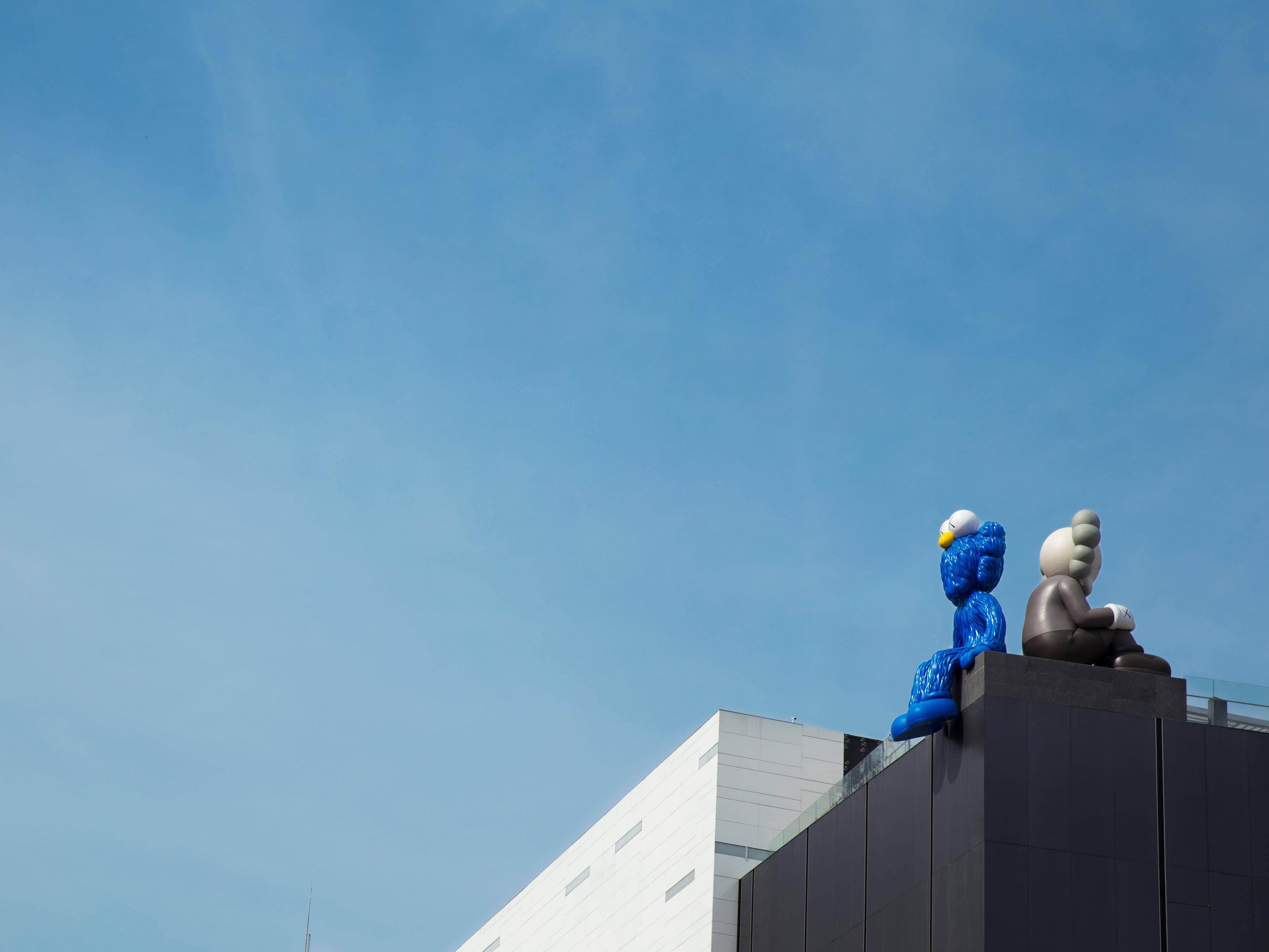 KAWS Seeing And Companion 壁紙 — 兩座大型風格化雕塑坐落在現代建築頂部，襯著清澈的藍天。