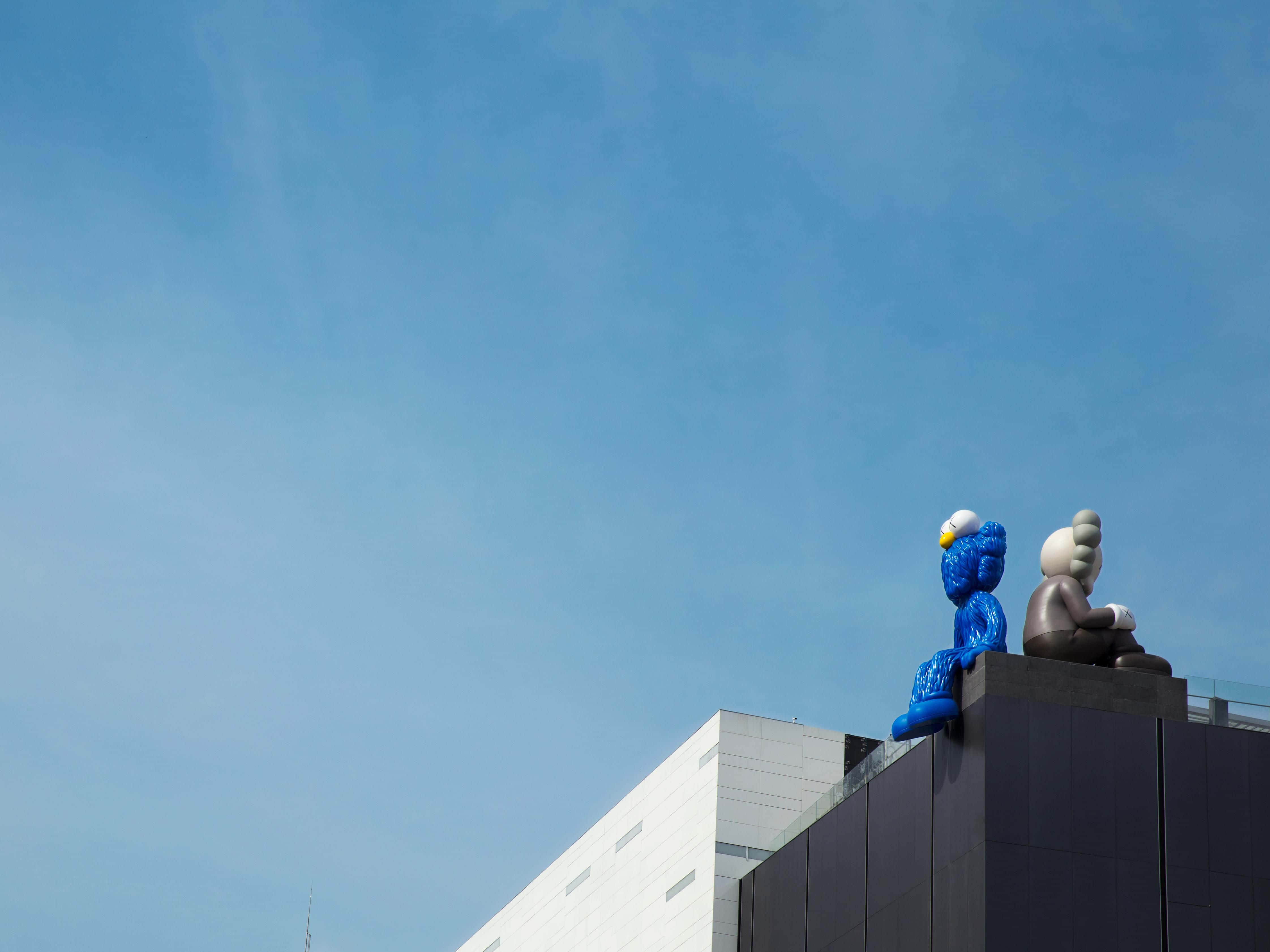 Kaws Bff 桌布 壁紙 — 兩座大型風格化雕塑坐落於現代建築頂部，背景是清澈的藍天。