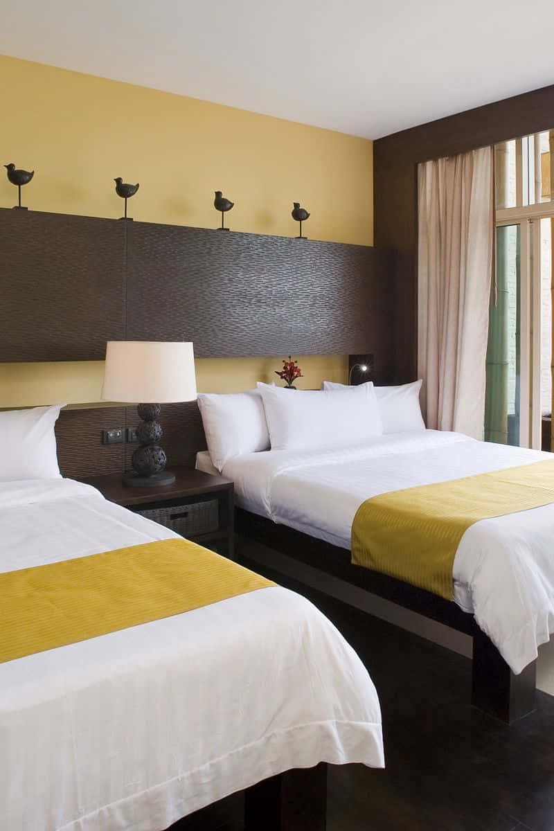 Hotel Room 桌布 壁紙 — 兩張整齊鋪好的床，配有白色床單和黃色點綴，位於一個帶有鳥類雕像的紋理床頭板下方。