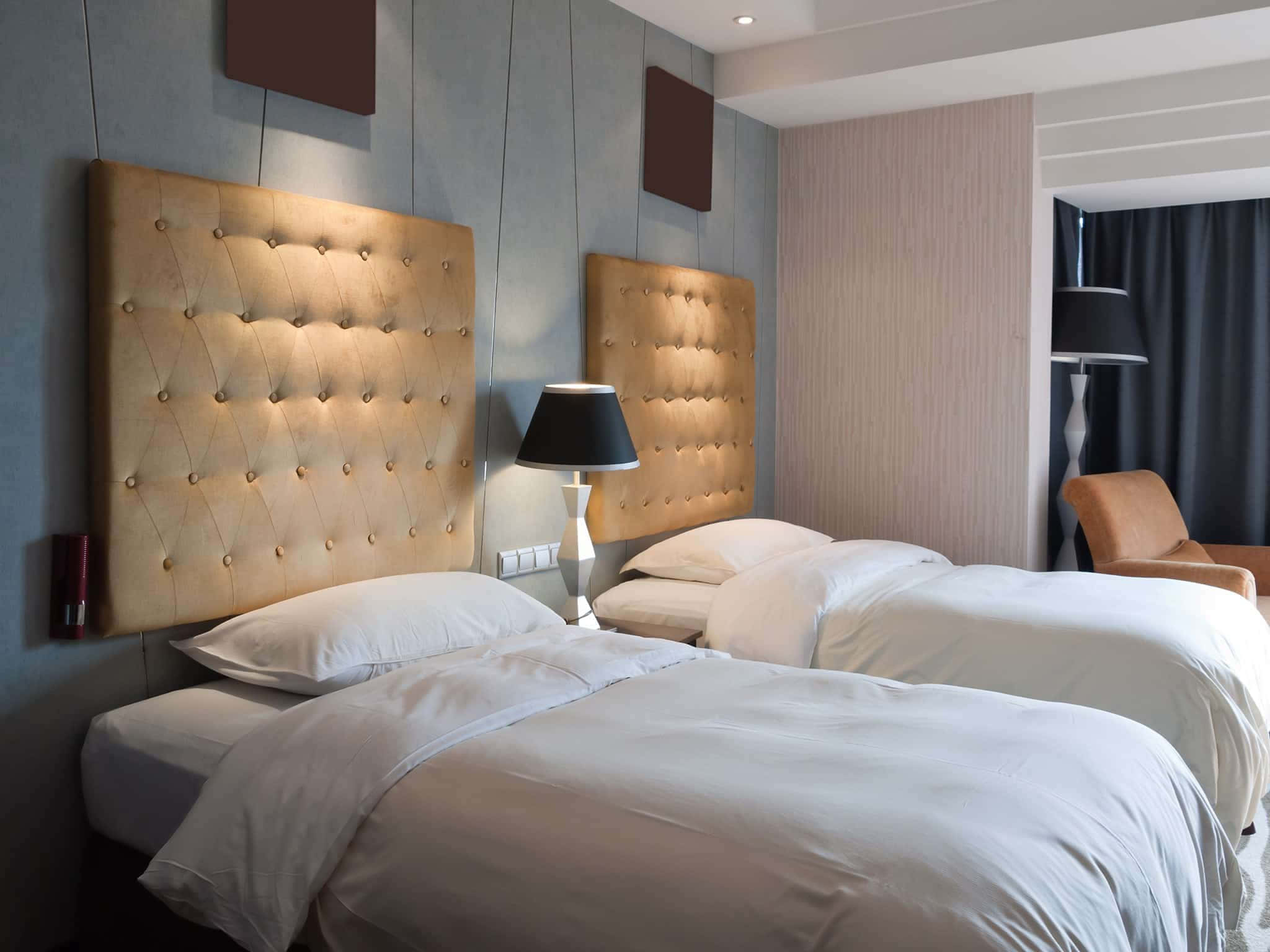 Hotel Room 桌布 壁紙 — 兩張整齊鋪好的床，配有簇絨米色床頭板，在現代酒店房間的環境照明下溫暖地發光。