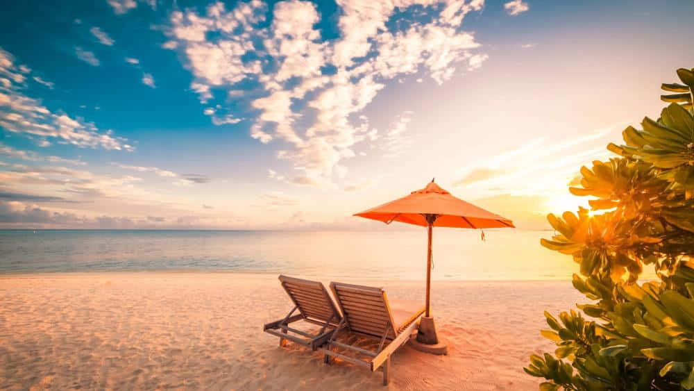 Beach Sunbed 桌布 壁紙 — 兩張木製躺椅在橙色遮陽傘下，金色沙灘上，日落時分，碧綠海洋與零星雲朵。