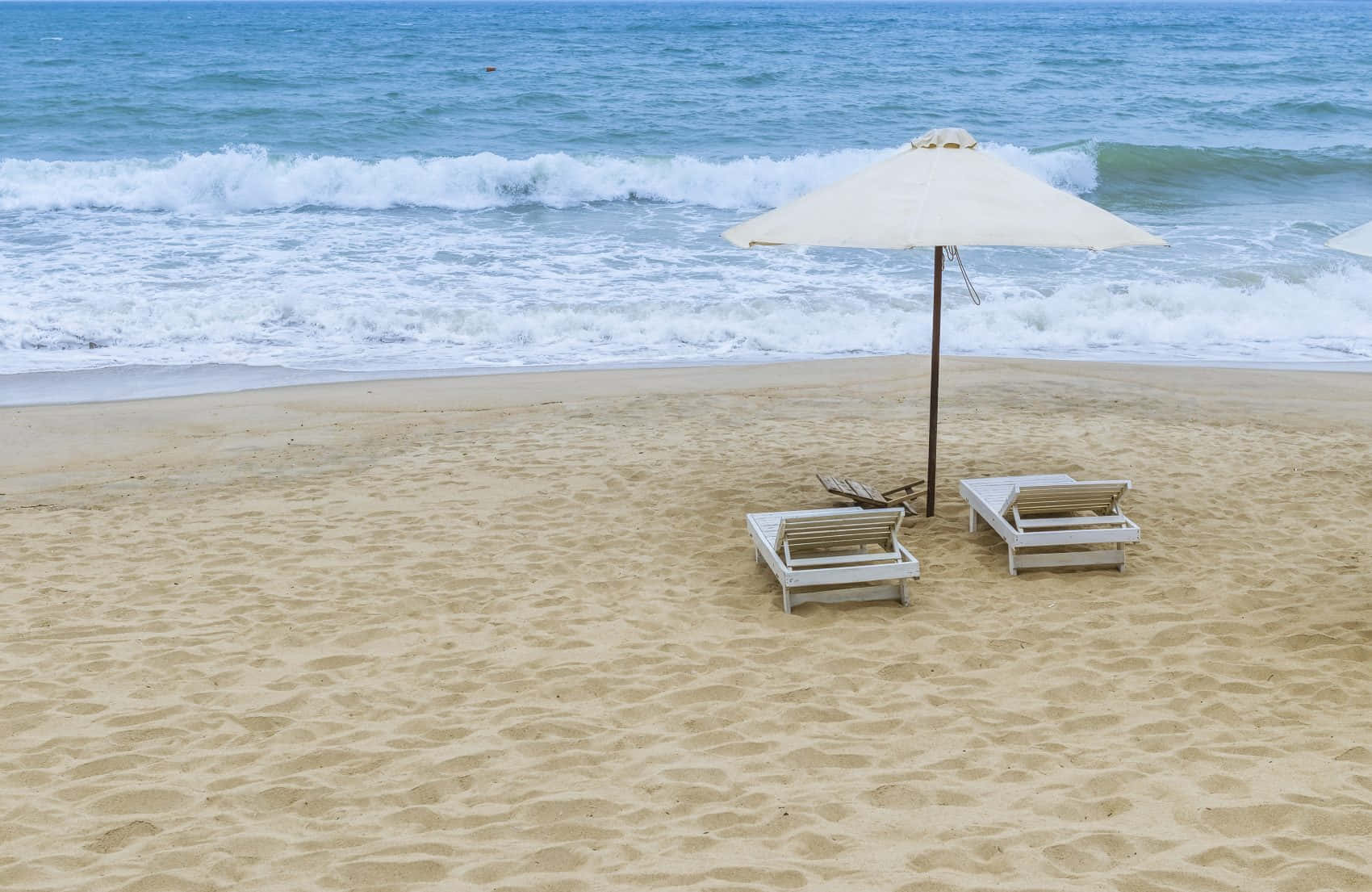Beach Sunbed 桌布 壁紙 — 兩張白色沙灘椅放在沙灘上的米色太陽傘下，背景中海浪拍打。