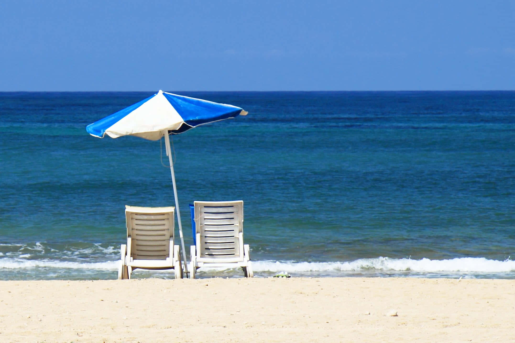 Beach Daybed 桌布 壁紙 — 兩張白色沙灘椅放在藍白相間的遮陽傘下，面向碧綠的海浪，位於沙灘上。