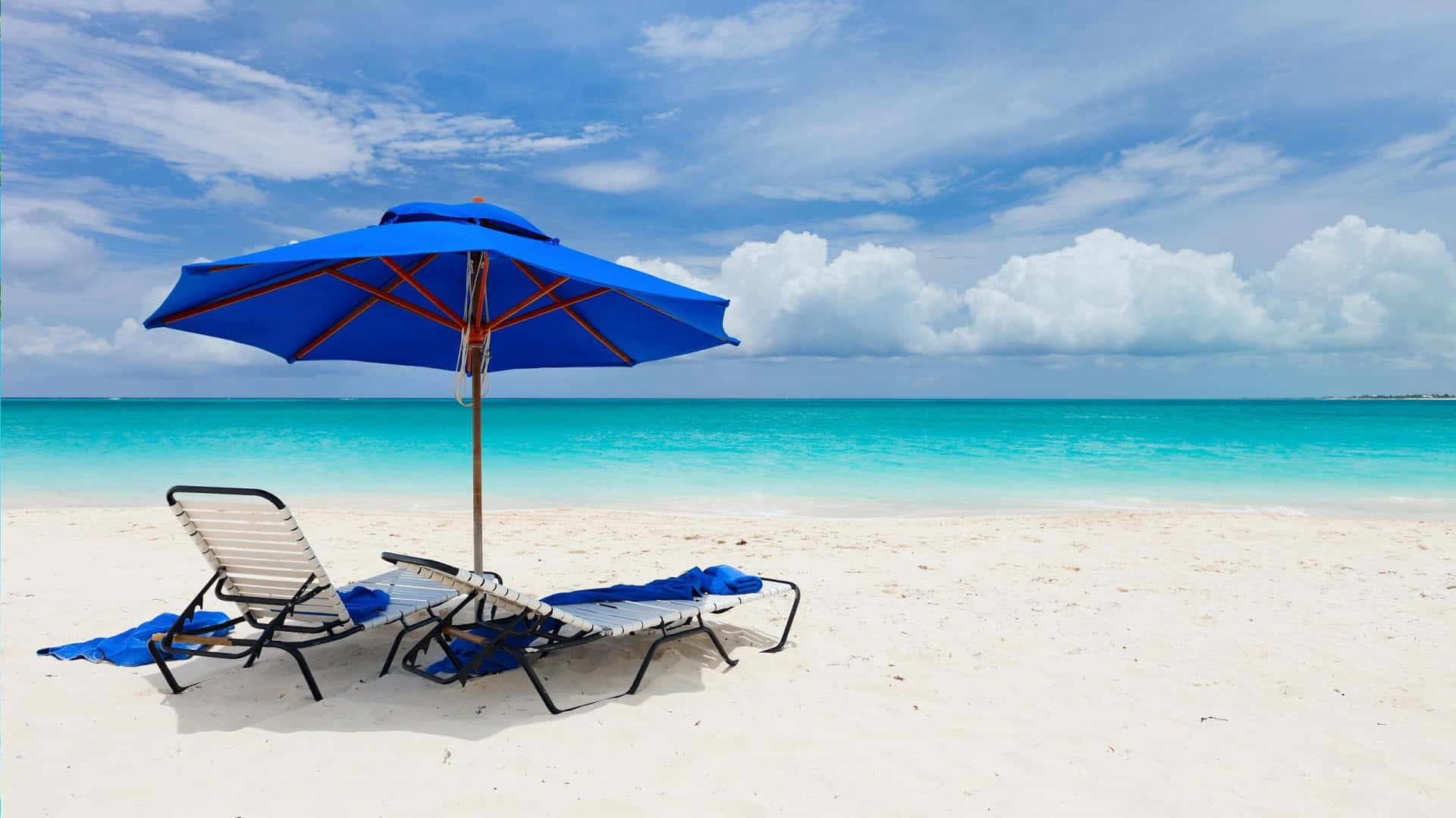 Beach Sunbed 桌布 壁紙 — 兩張躺椅在明亮的藍色遮陽傘下，面向碧綠海洋和多雲天空的純白沙灘。