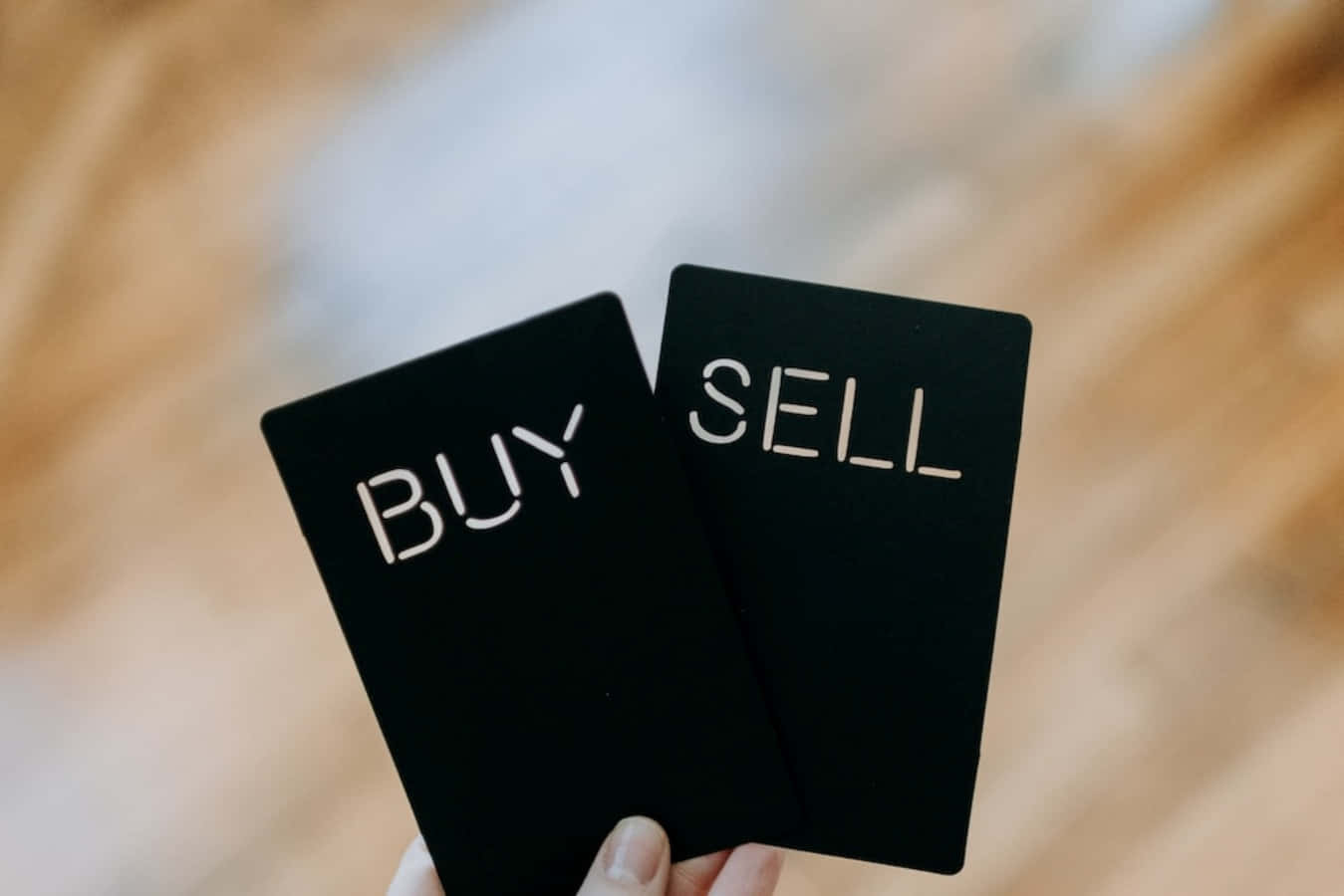 buy 桌布 壁紙 — 兩張黑色卡片，上面有白色文字寫著「BUY」和「SELL」，握在手中，背景是模糊的木地板。