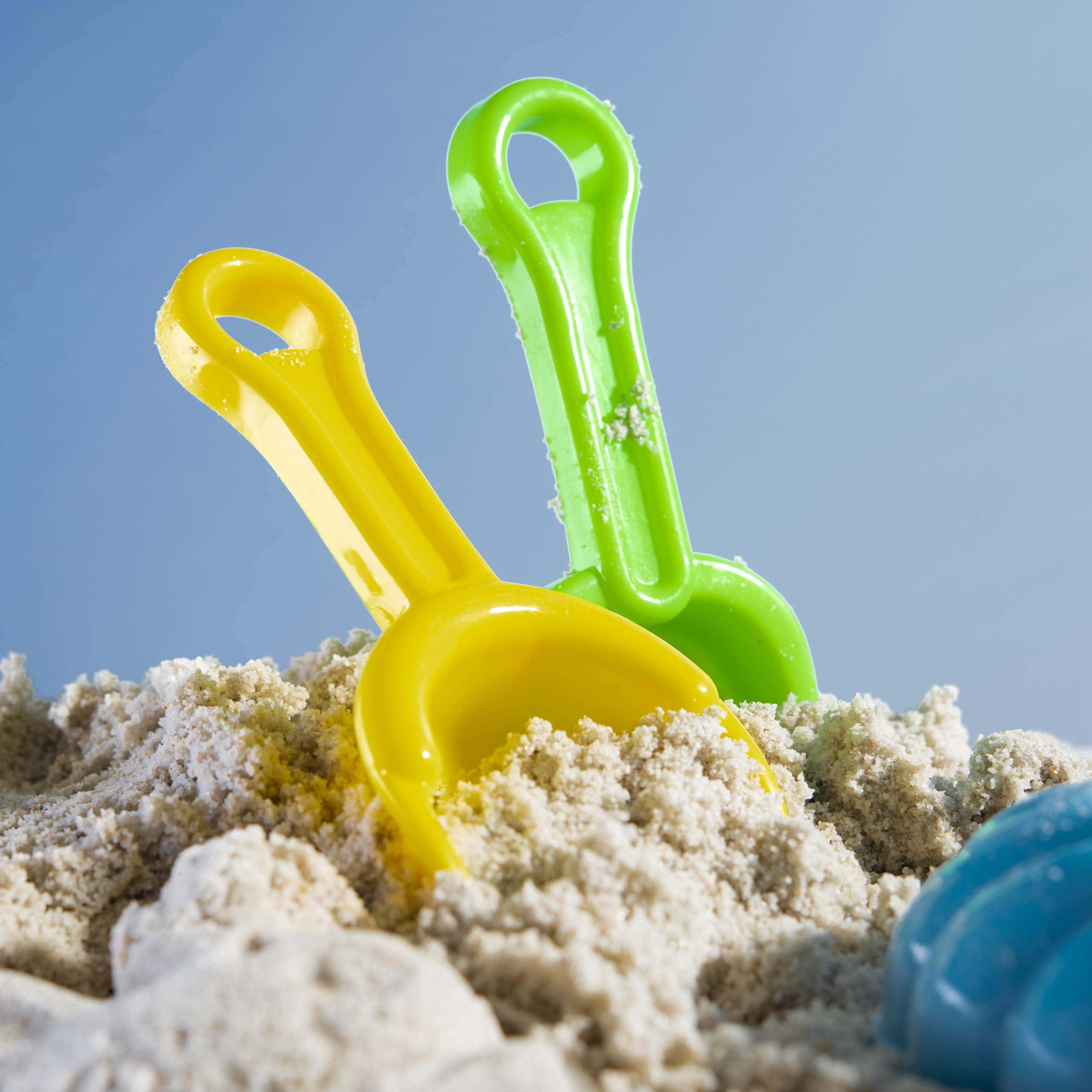 Beach Toys 桌布 壁紙 — 兩把鮮黃色和綠色的塑膠沙灘鏟從有紋理的沙中伸出，映襯著清澈的藍天。