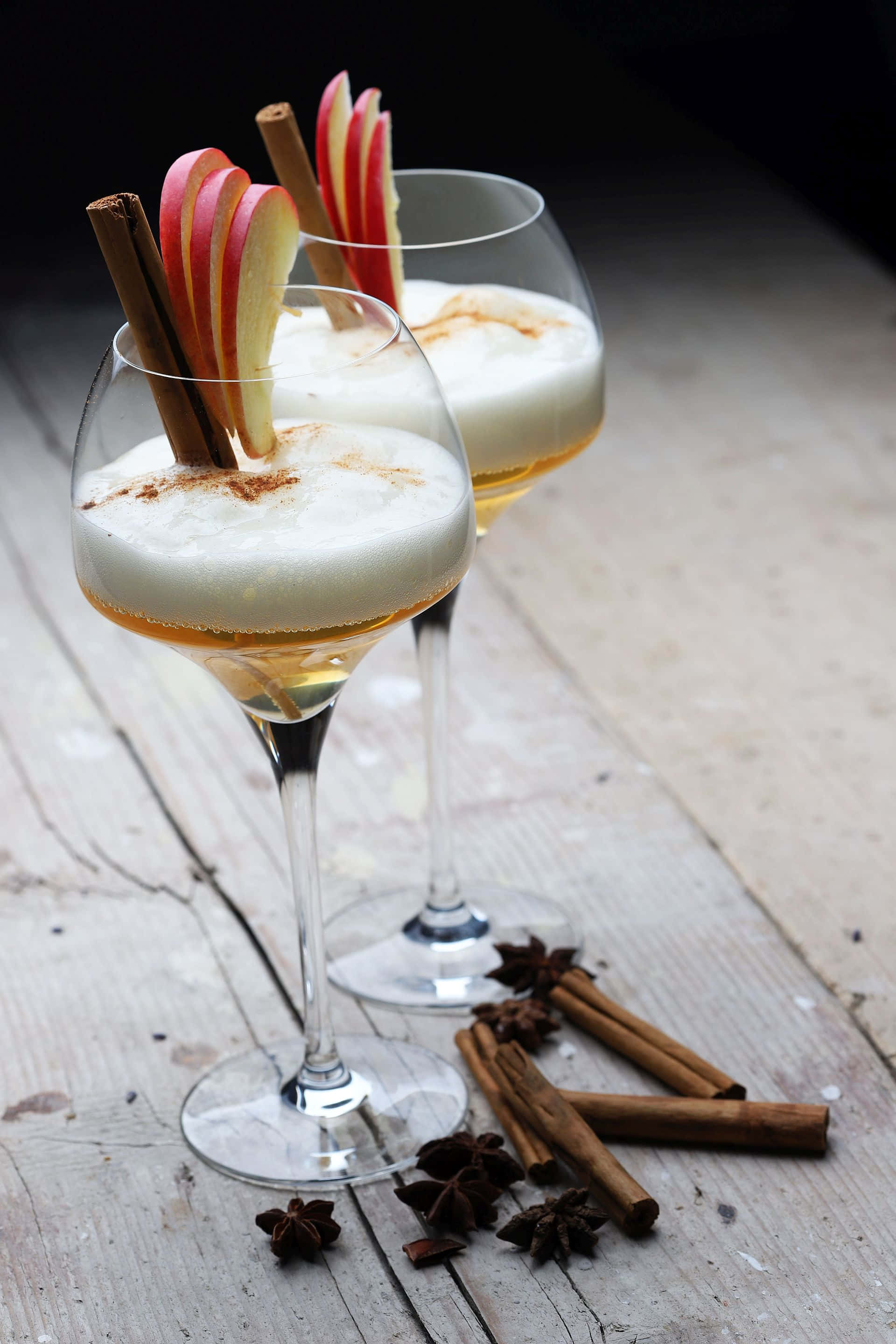 Winter Drinks 桌布 壁紙 — 兩杯優雅的雞尾酒，上面有泡沫，並以蘋果片和肉桂棒裝飾，放在質樸的木質表面上。
