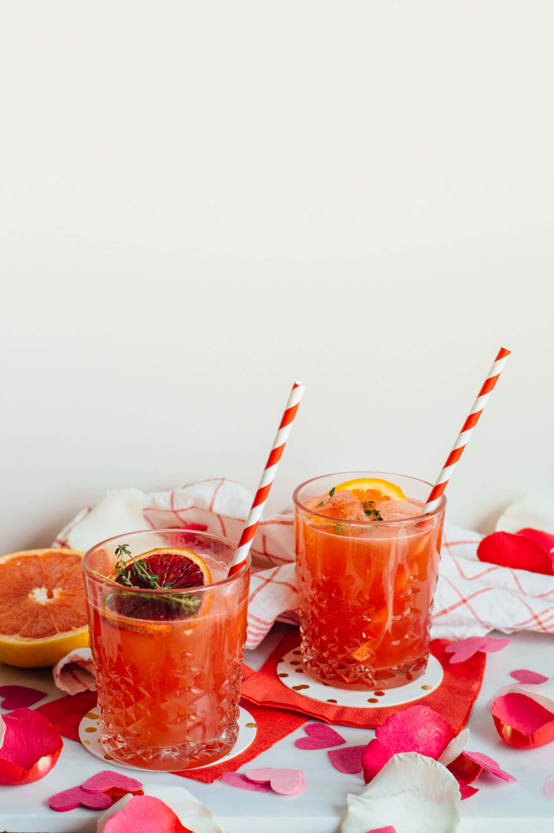 Winter Drinks 桌布 壁紙 — 兩杯充滿活力的粉紅色柑橘飲品，配有條紋吸管，玫瑰花瓣散落在紅色心形圖案的布上。