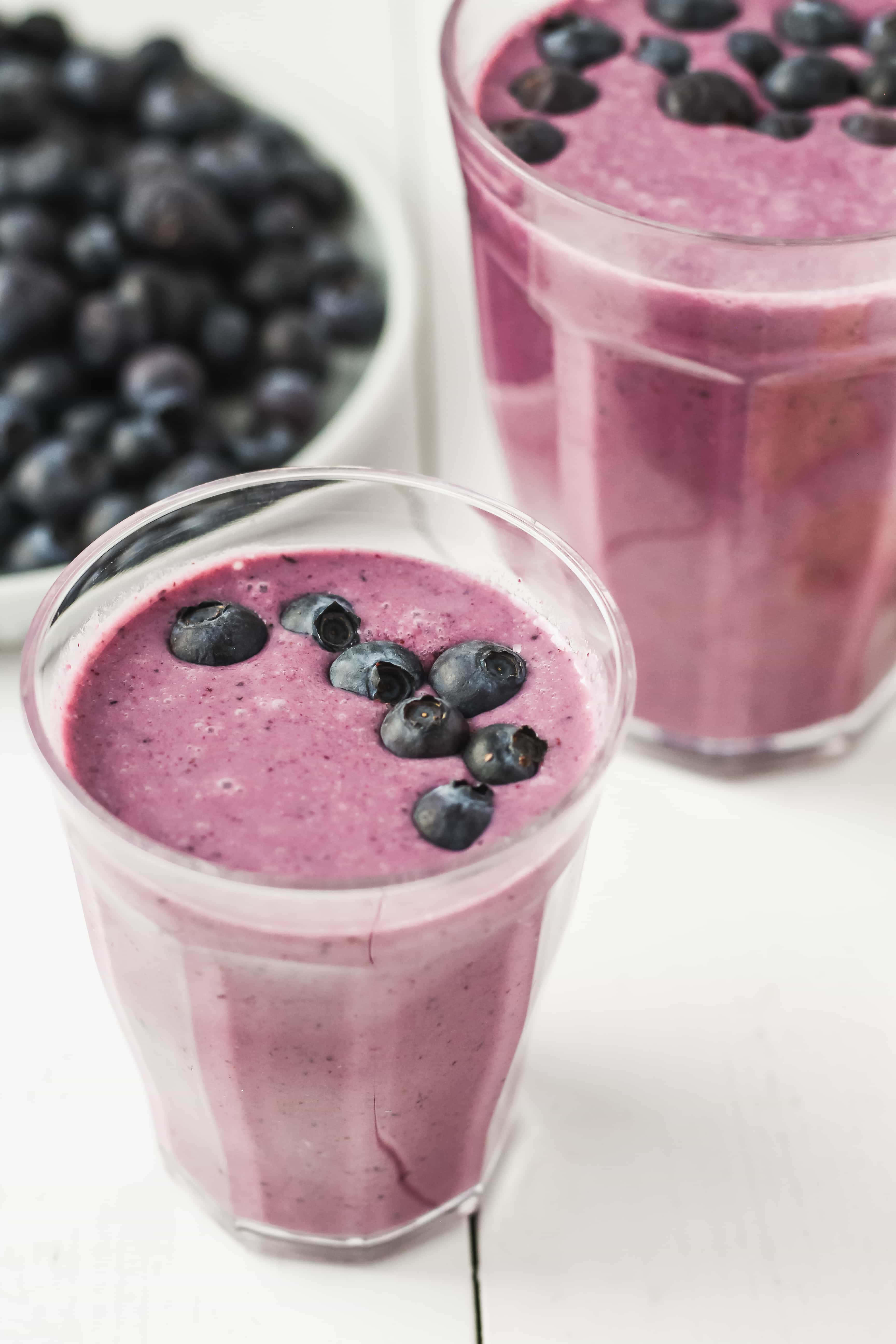 Blueberry Smoothie 桌布 壁紙 — 兩杯充滿活力的紫色藍莓冰沙，上面放上新鮮藍莓，旁邊放著一碗漿果，放在白色木質表面上。