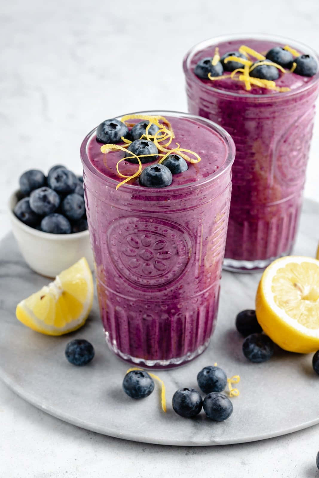 Blueberry Smoothie 桌布 壁紙 — 兩杯充滿活力的紫色藍莓檸檬冰沙，盛載於質感玻璃杯中，以新鮮藍莓和檸檬皮裝飾，置於大理石表面上。