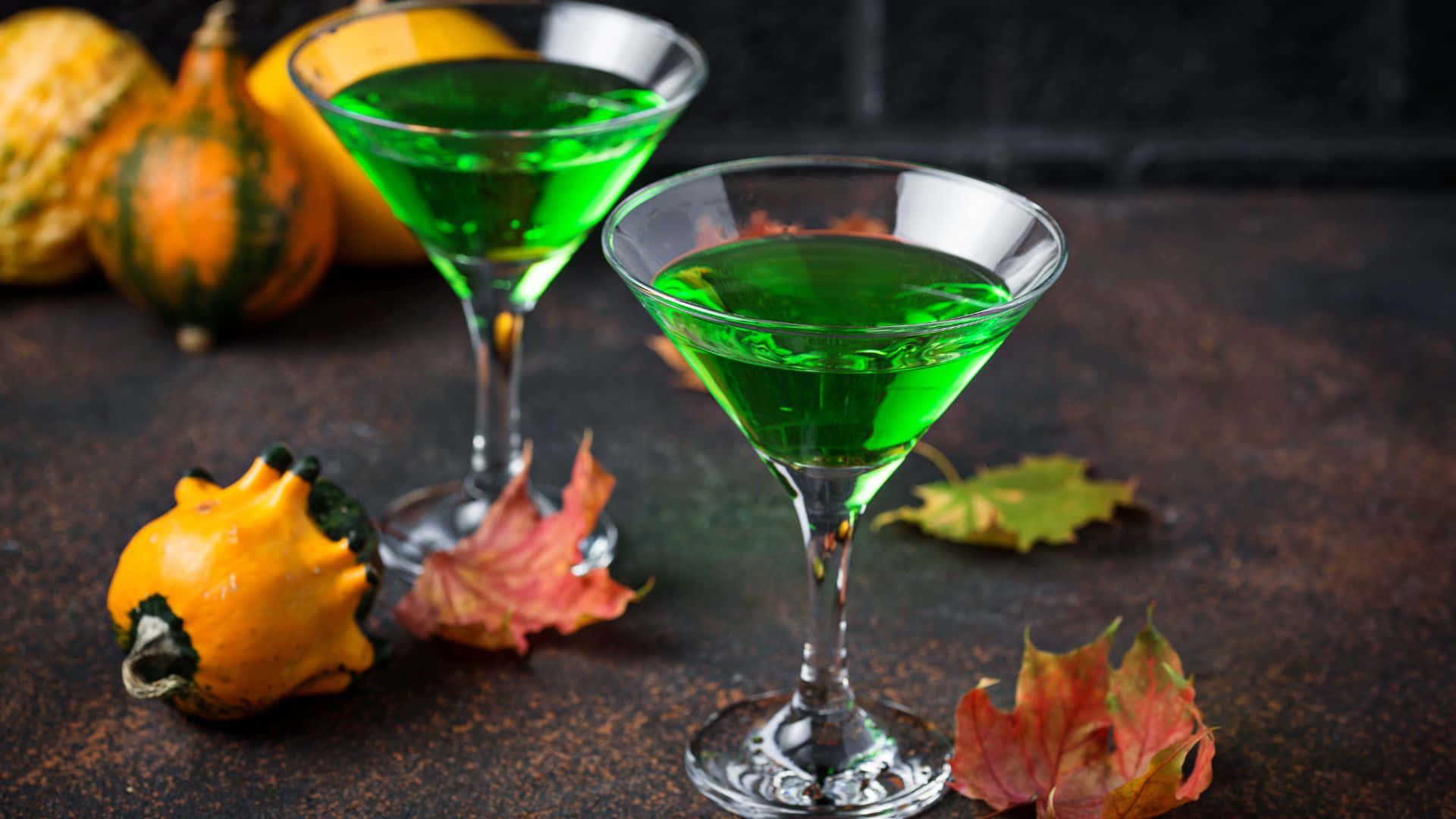 Spooky Cocktails 桌布 壁紙 — 兩杯充滿活力的綠色雞尾酒盛載於馬天尼杯中，周圍環繞著秋季葫蘆和色彩繽紛的樹葉，置於鄉村風格的表面上。