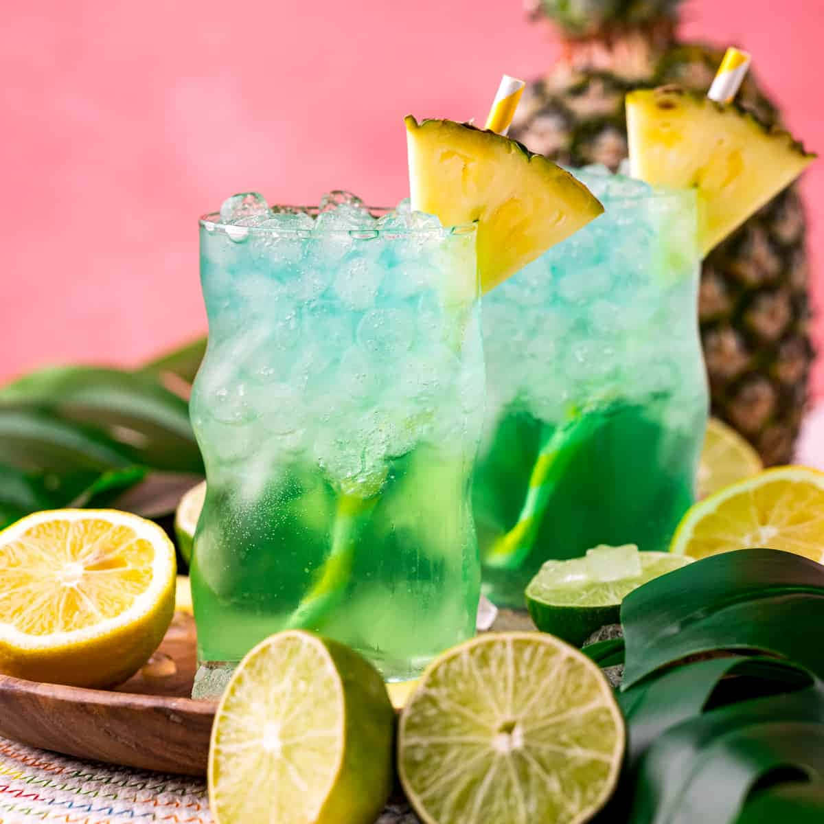 Beautiful Drink 桌布 壁紙 — 兩杯充滿活力的藍綠色熱帶雞尾酒，搭配菠蘿裝飾和柑橘片，放在粉紅色背景前的木托盤上。