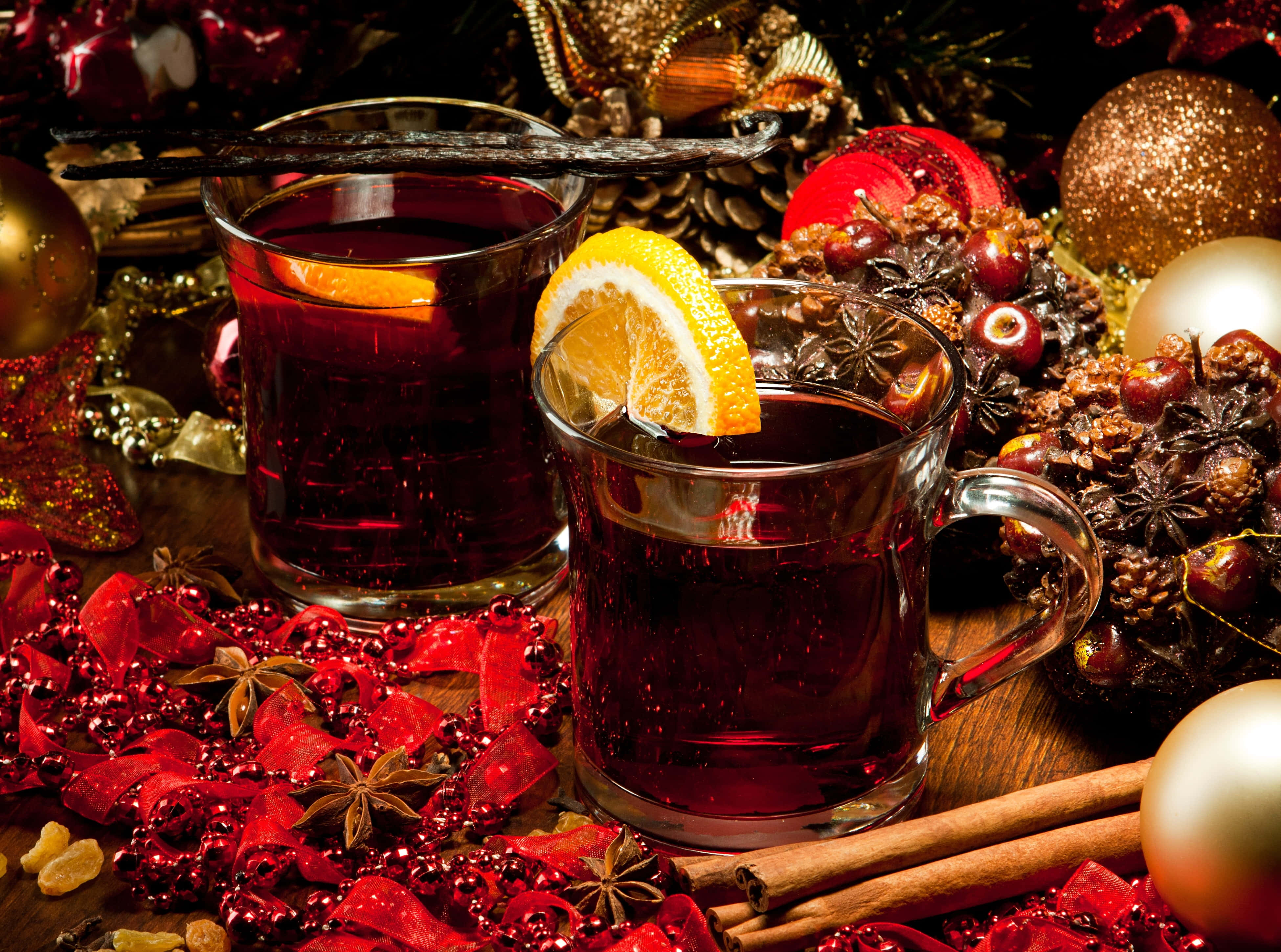 Mulled Wine 桌布 壁紙 — 兩杯冒著熱氣的香料熱紅酒，飾以柑橘和雲呢拿，放在木質表面上，周圍是節日紅色裝飾品、松果和香料。