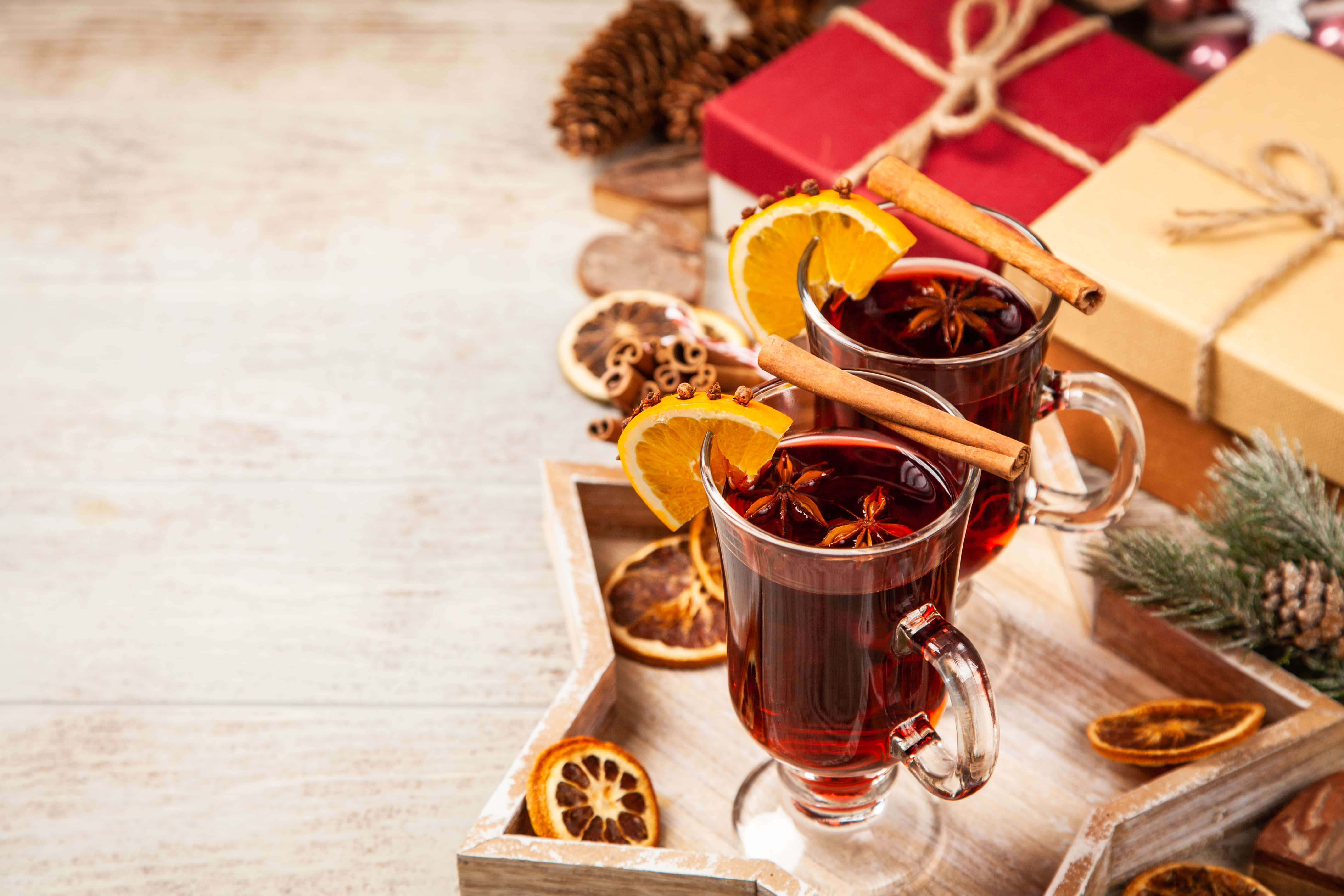 Mulled Wine 桌布 壁紙 — 兩杯冒著熱氣的香料熱紅酒，飾以橙片、肉桂棒和八角，放在木托盤上，旁邊有節日禮物、松果和乾柑橘。