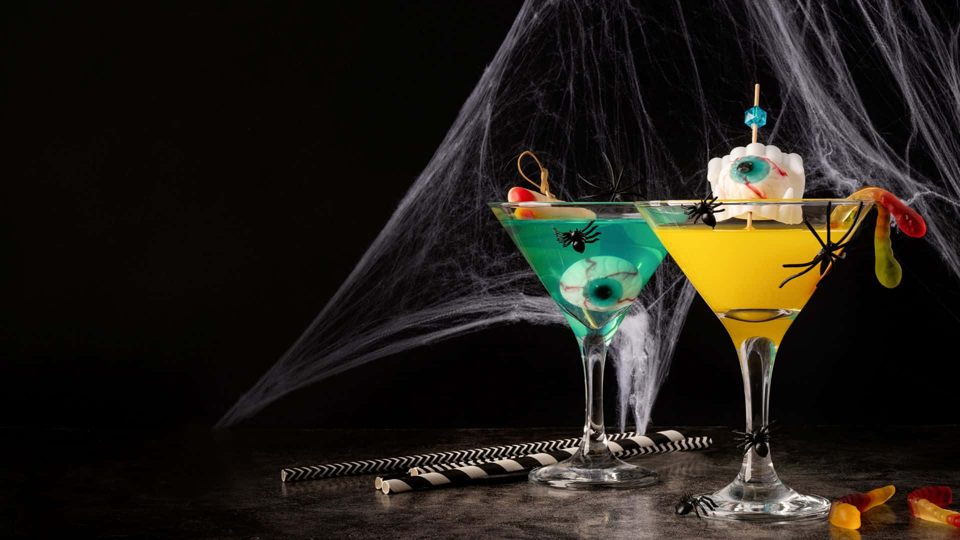 Spooky Cocktails 桌布 壁紙 — 兩杯恐怖萬聖節雞尾酒，盛載於馬提尼杯中，飾以眼珠和蜘蛛，背景為黑色帶蜘蛛網。