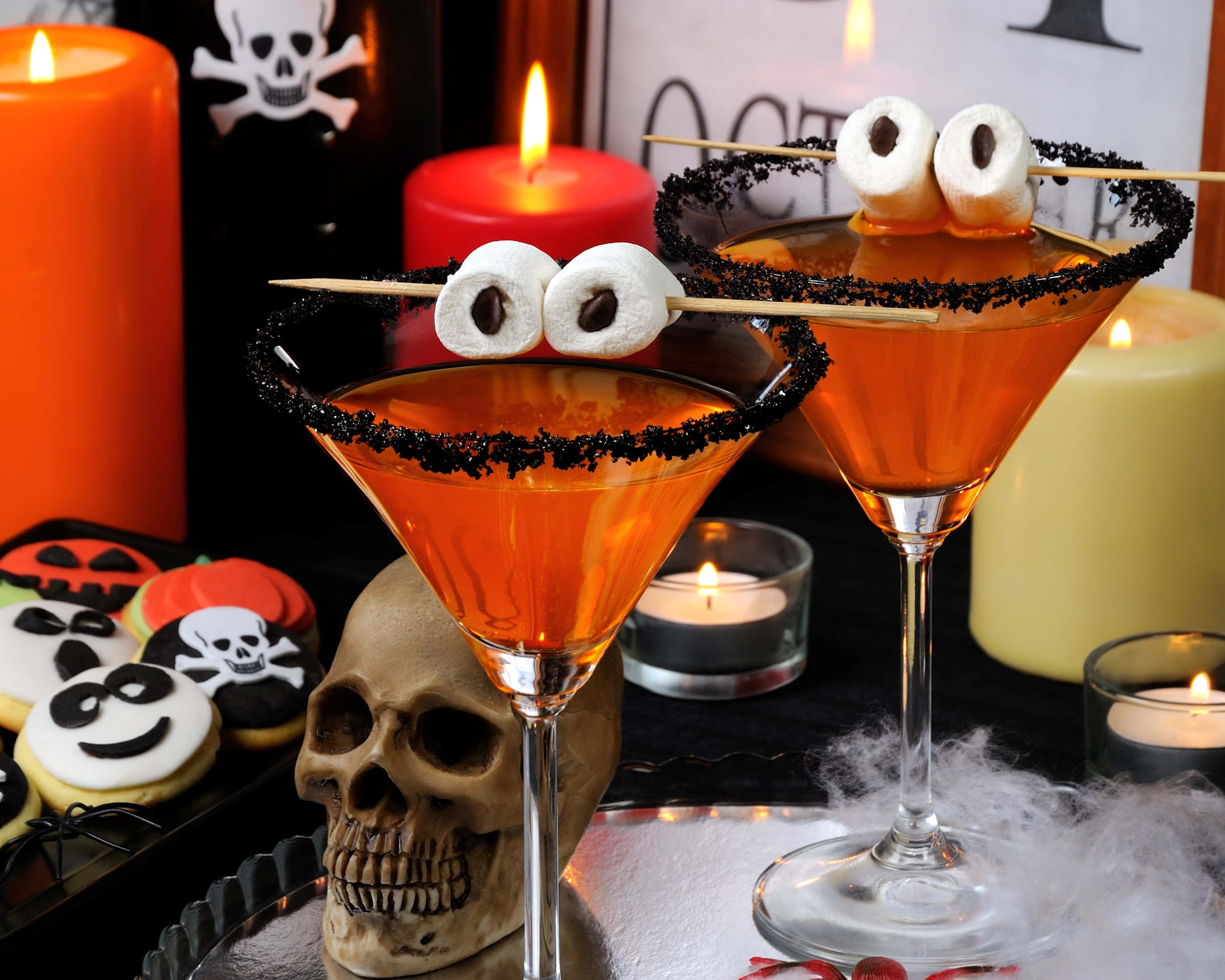 Halloween Cocktails 桌布 壁紙 — 兩杯橙色萬聖節雞尾酒，帶有棉花糖眼睛和黑色糖邊，旁邊放著一個骷髏頭和發光的蠟燭。