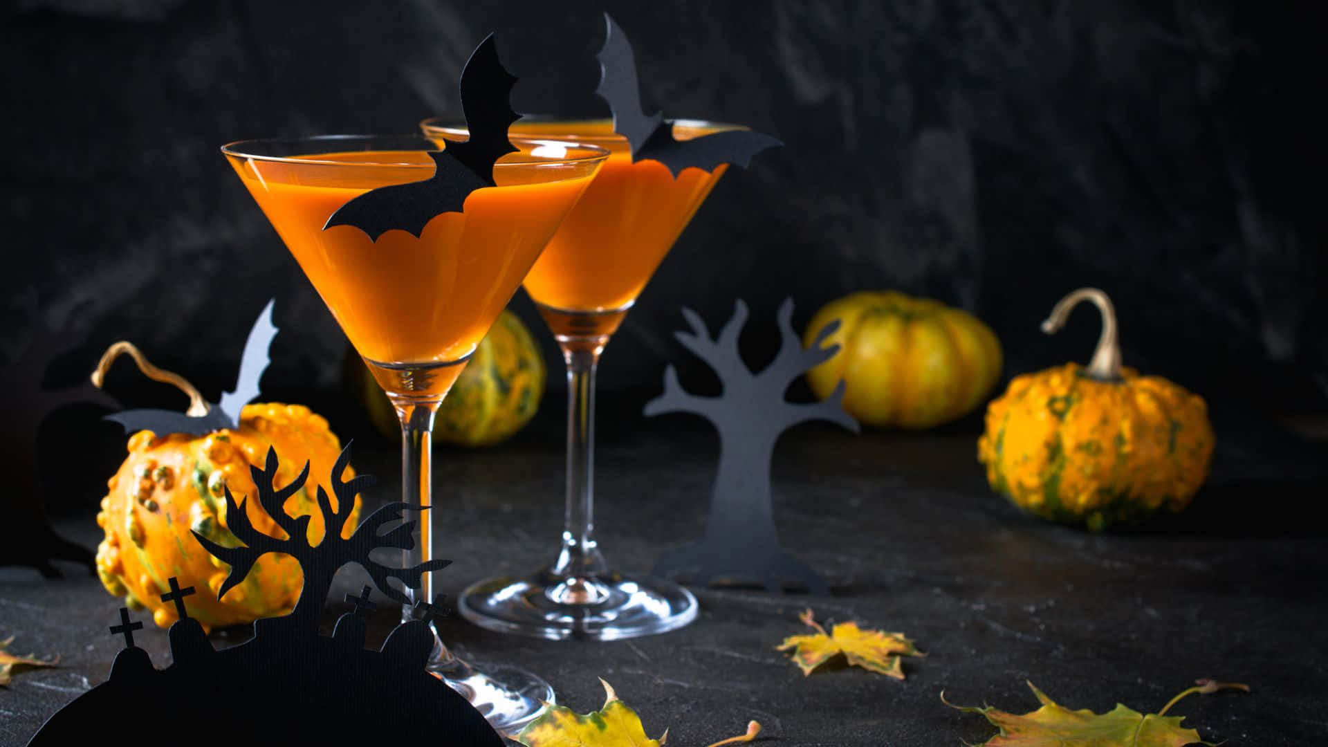 Spooky Cocktails 桌布 壁紙 — 兩杯橙色萬聖節雞尾酒，飾有蝙蝠剪影，旁邊有南瓜和恐怖剪紙，放在深色石面上。