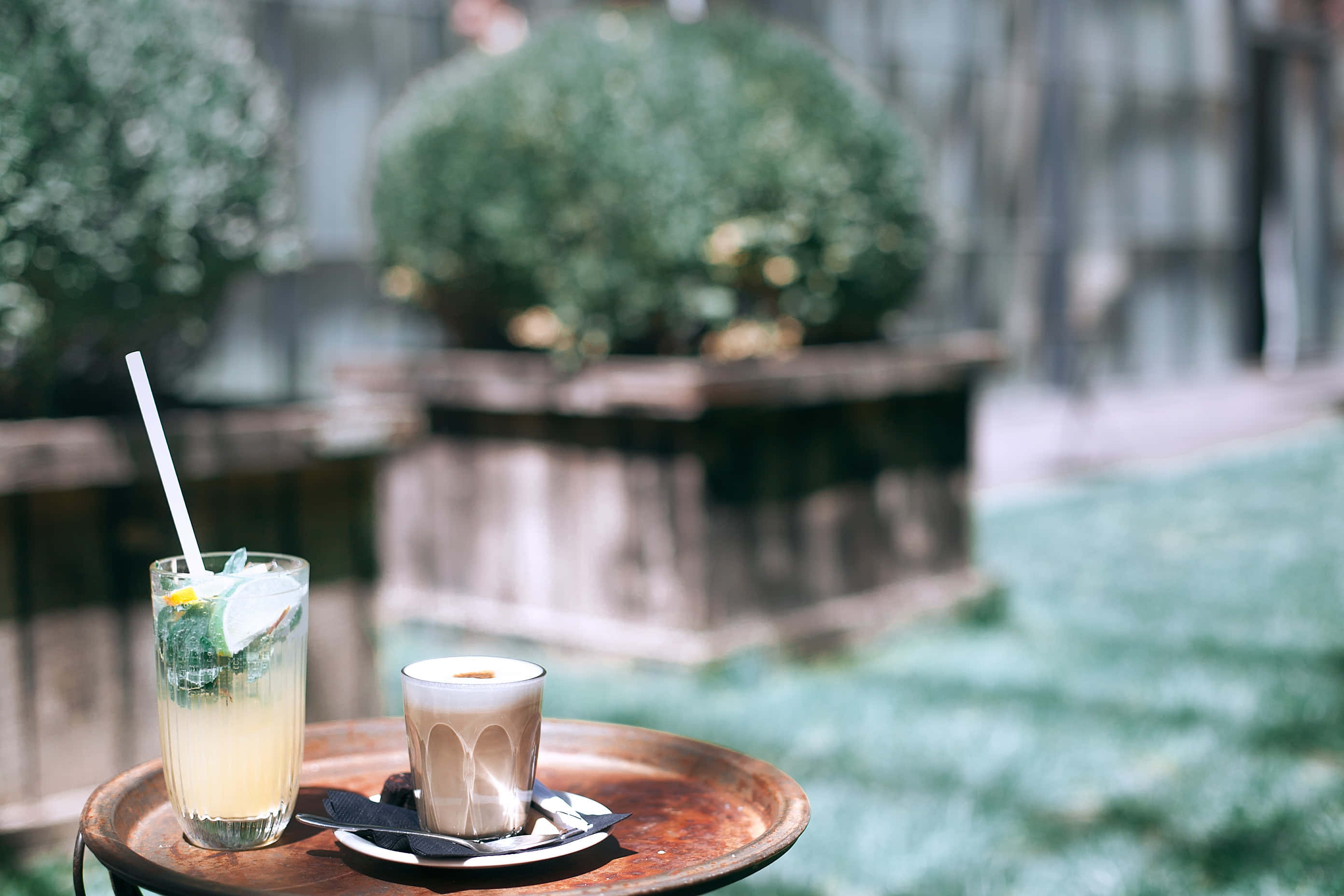 Summer Drinks 桌布 壁紙 — 兩杯清涼飲品放在戶外一個質樸的木托盤上，背景是模糊的綠植和現代建築，在柔和日光下。