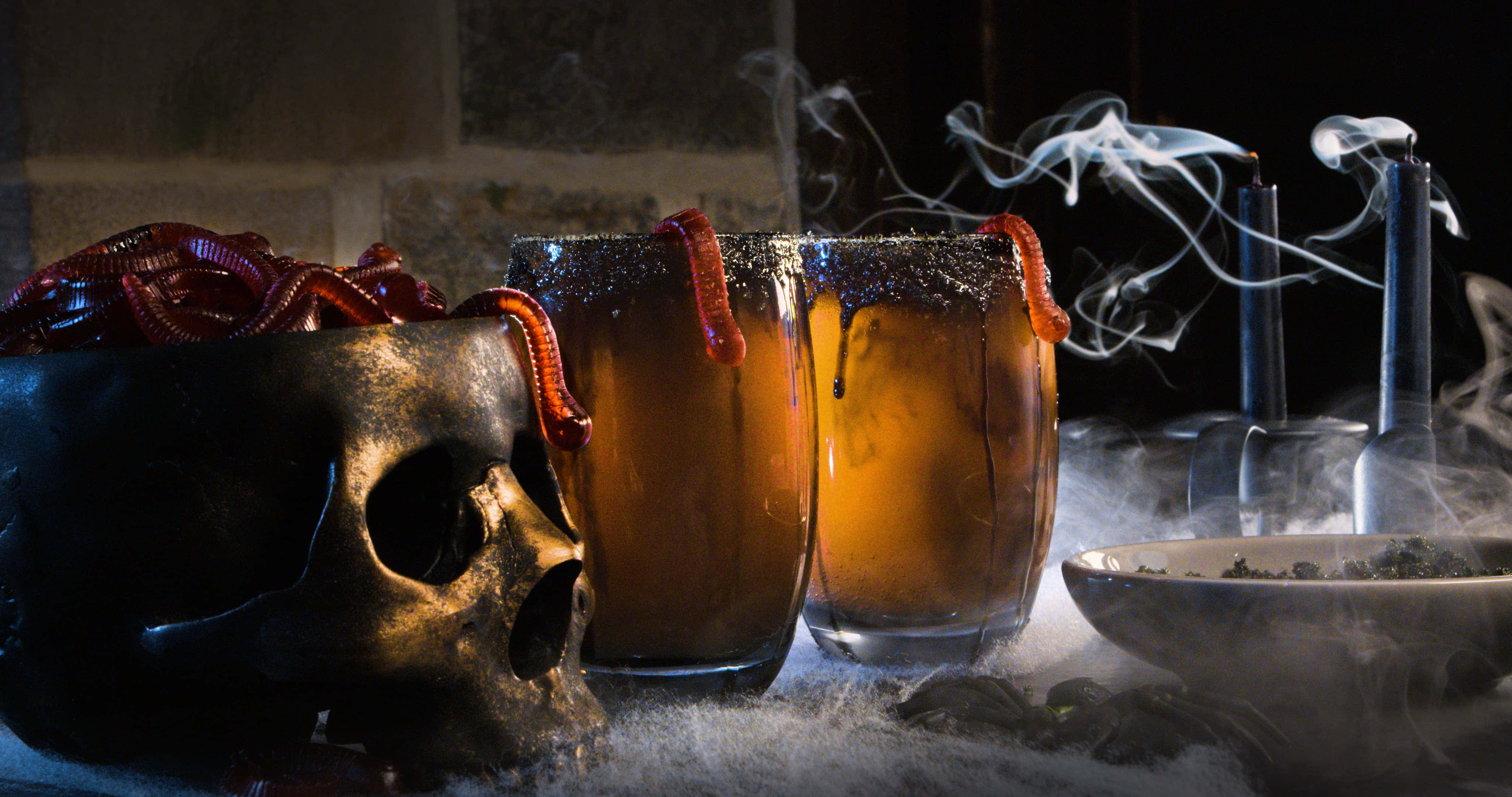 Spooky Cocktails 桌布 壁紙 — 兩杯煙燻琥珀色飲品，內有蠕動的蟲子，旁邊放著一個骷髏頭和冒煙的煙斗，置於結霜的表面上。