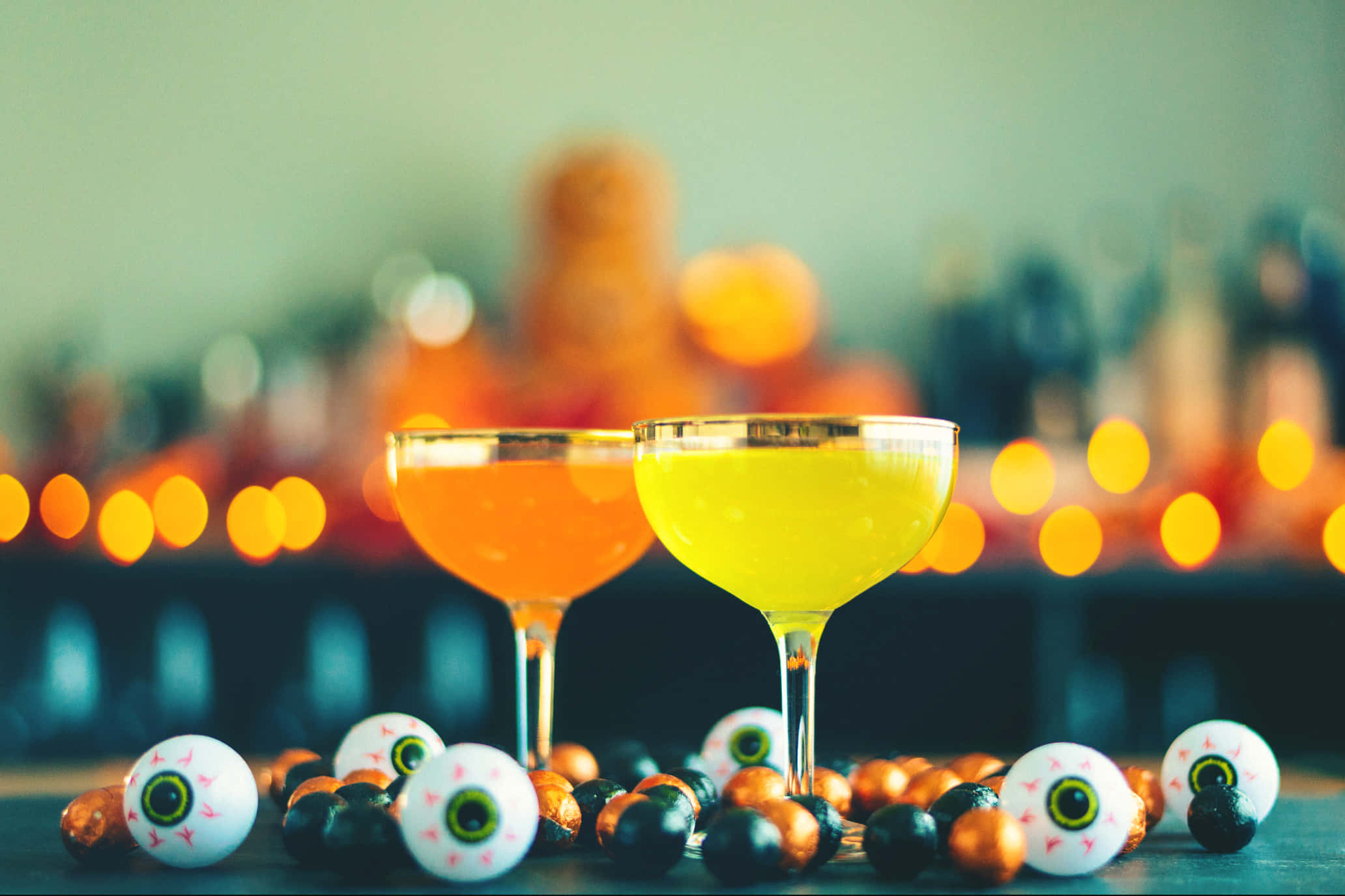 Spooky Cocktails 桌布 壁紙 — 兩杯鮮艷的橙色和黃色雞尾酒放在糖果眼珠和朱古力旁邊，背景是模糊的萬聖節主題酒吧。