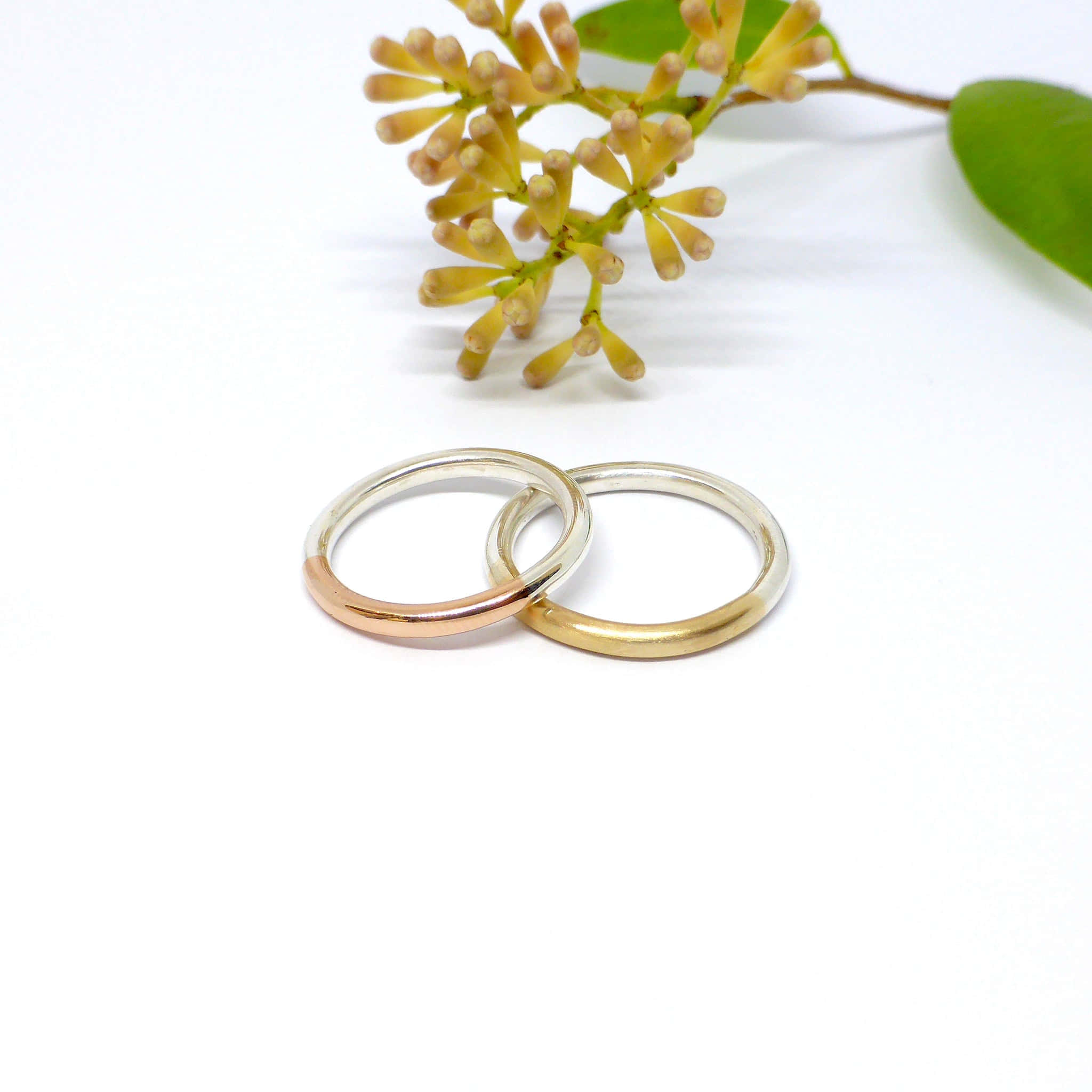 Wedding Rings 桌布 壁紙 — 兩枚交織的結婚戒指，一枚玫瑰金一枚黃金，放在純白表面上，旁邊是精緻的黃色花朵。