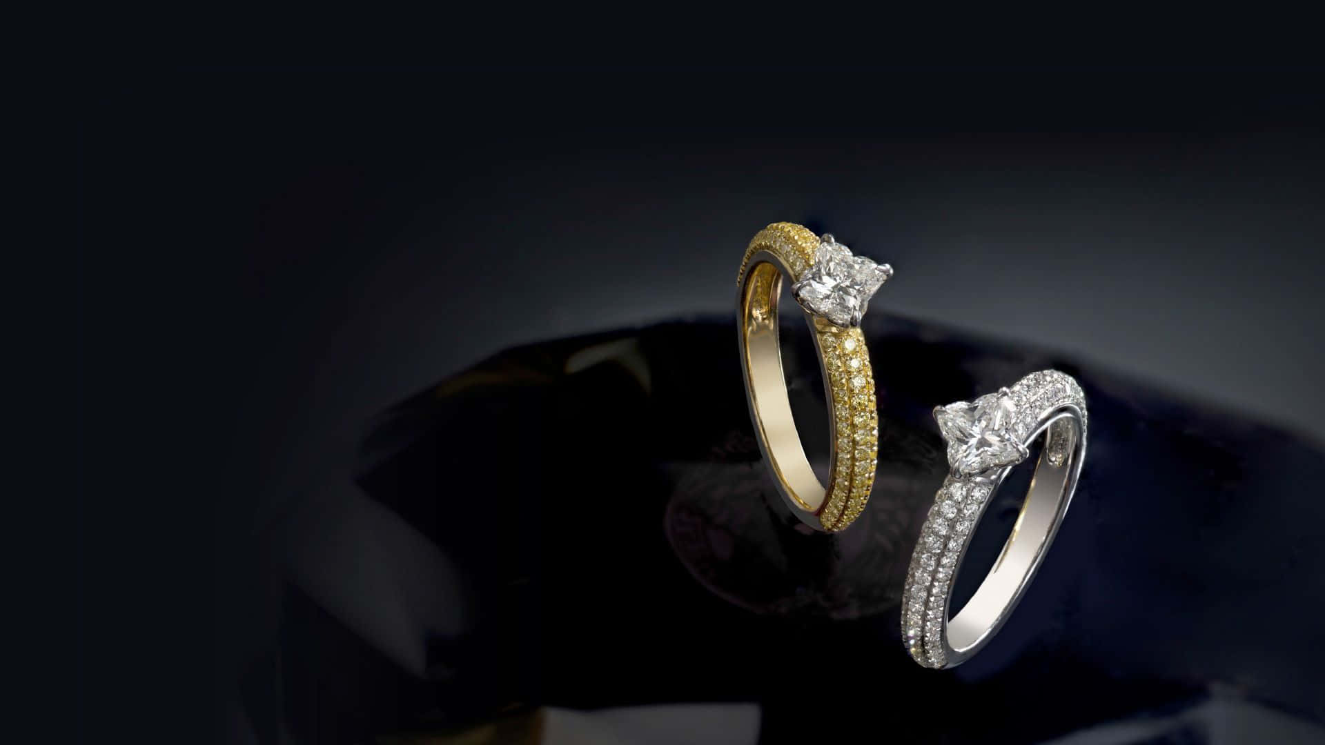 Gold Jewellery 桌布 壁紙 — 兩枚優雅的鑽石戒指，一枚黃金一枚白金，在深色反光表面上閃耀。