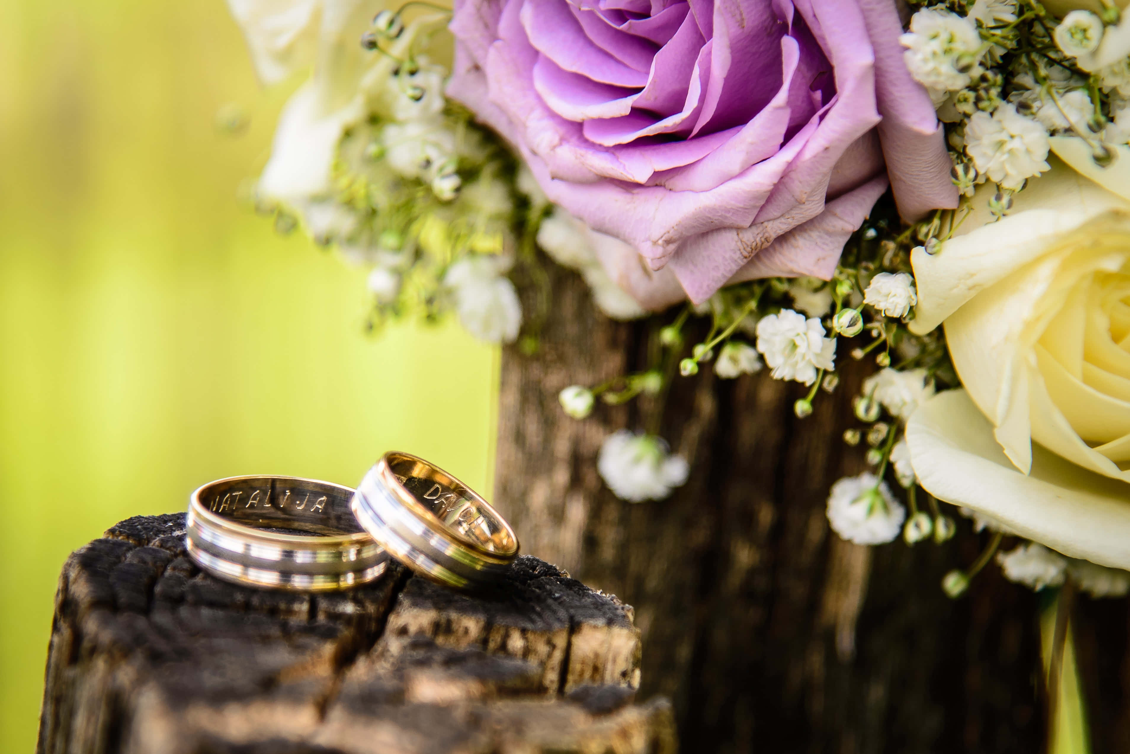 Wedding Rings 桌布 壁紙 — 兩枚刻有圖案的結婚戒指放在一個風化的樹樁上，旁邊是一束紫色和白色的玫瑰。