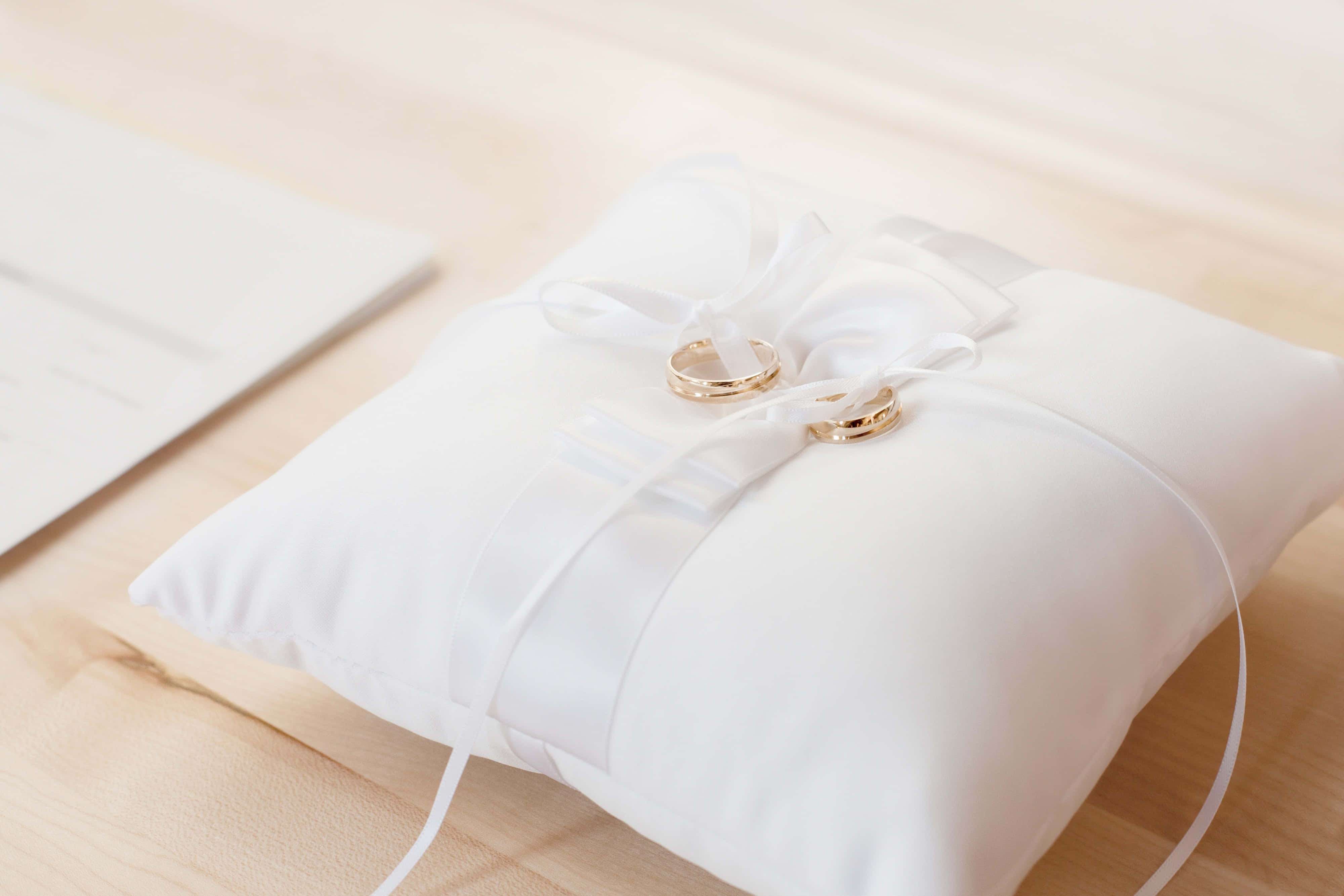 Wedding Rings 桌布 壁紙 — 兩枚金結婚戒指放在一個綁有蝴蝶結的白色緞面枕頭上，置於淺色木質表面。