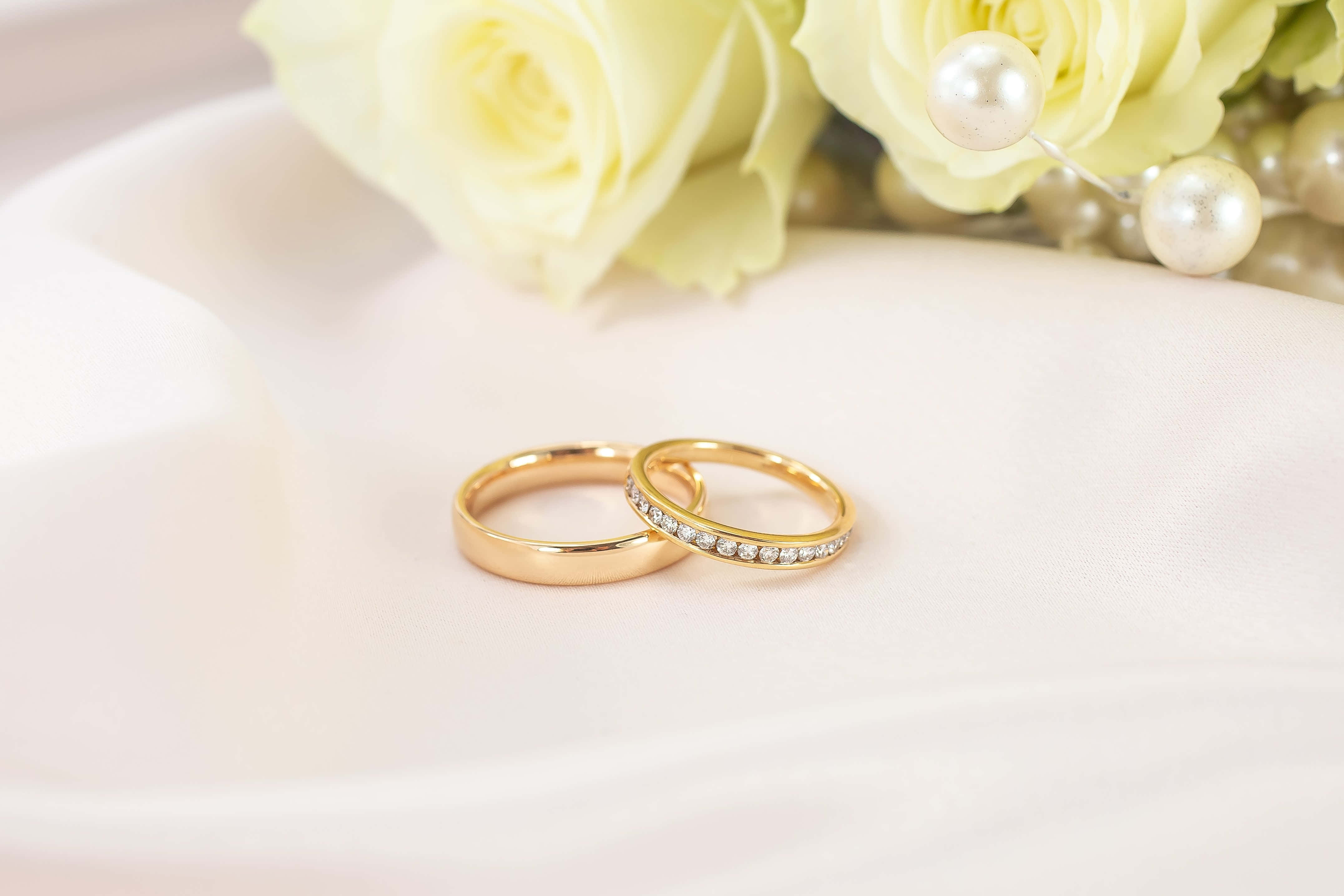 Wedding Rings 桌布 壁紙 — 兩枚金色結婚戒指放在柔軟的白色布料上，旁邊有淡黃色玫瑰和珍珠。