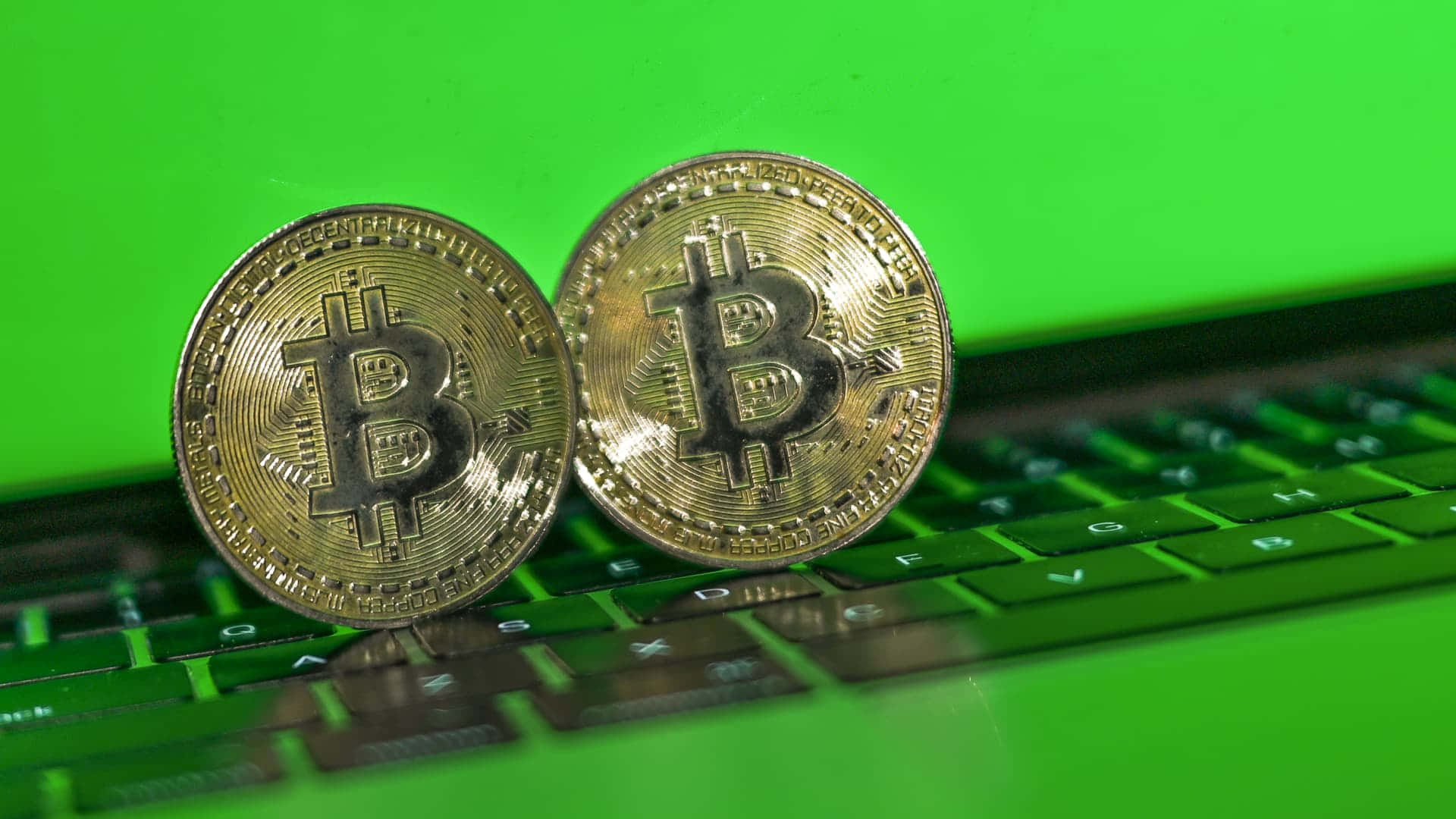 加密貨幣 桌布 壁紙 — 兩枚閃閃發光的金色比特幣硬幣放在一個充滿活力的綠色筆記型電腦鍵盤上，對焦清晰，景深較淺。