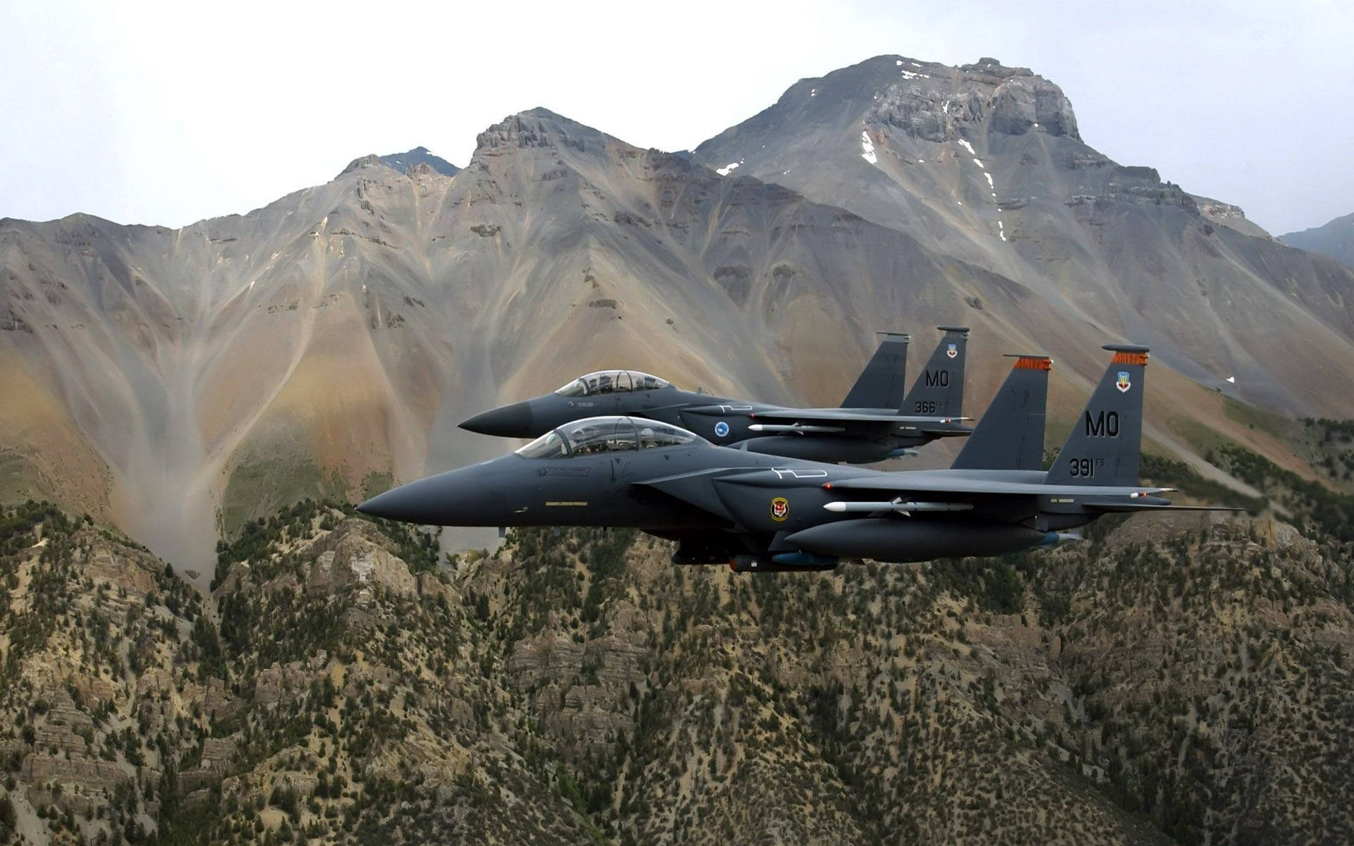 軍事動圖 桌布 壁紙 — 兩架戰鬥機在陰天嘅天空下，喺崎嶇嘅雪山上面編隊飛行。