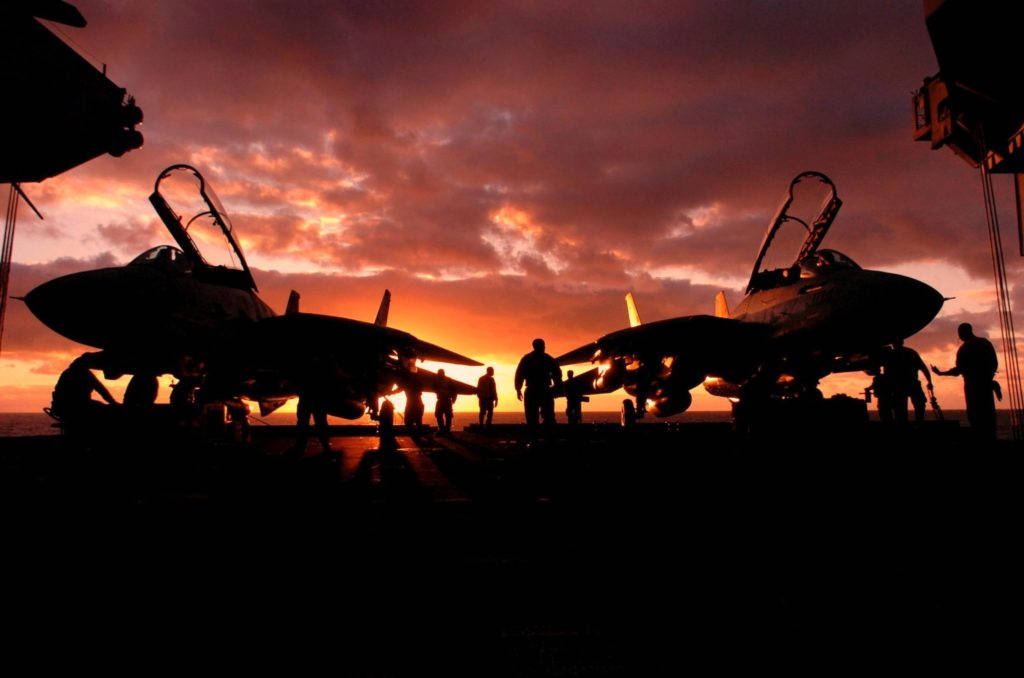 兩架美國海軍戰機停泊於航空母艦甲板 壁紙 — 喺熾熱嘅日落天空背景下，航空母艦甲板上嘅戰鬥機同機組人員嘅剪影。