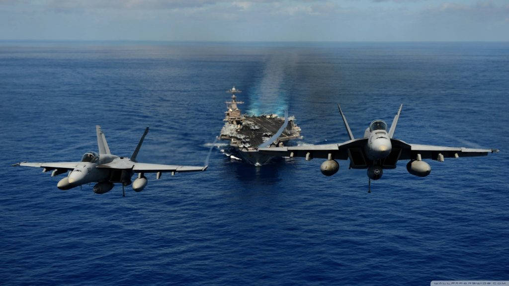 兩架美國海軍戰機起飛 壁紙 — 兩架戰鬥機編隊飛行在廣闊的藍色海洋上空，遠處有一艘航空母艦拖著尾跡。